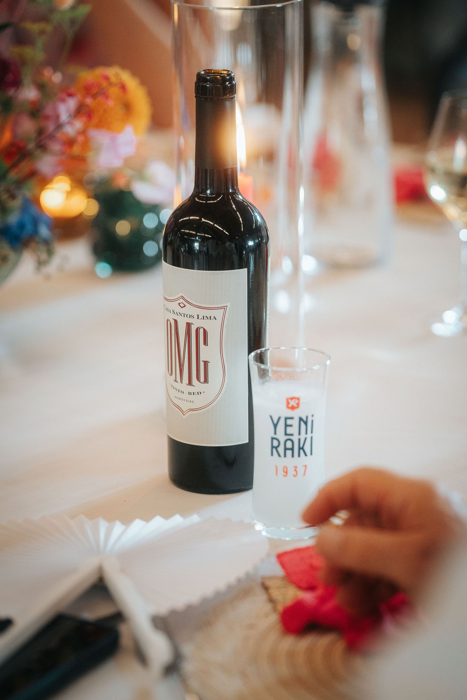 Bottle of OMG red wine beside a Yeni Raki 1937 glass on a festive table with flowers, candles, and folded paper fans.