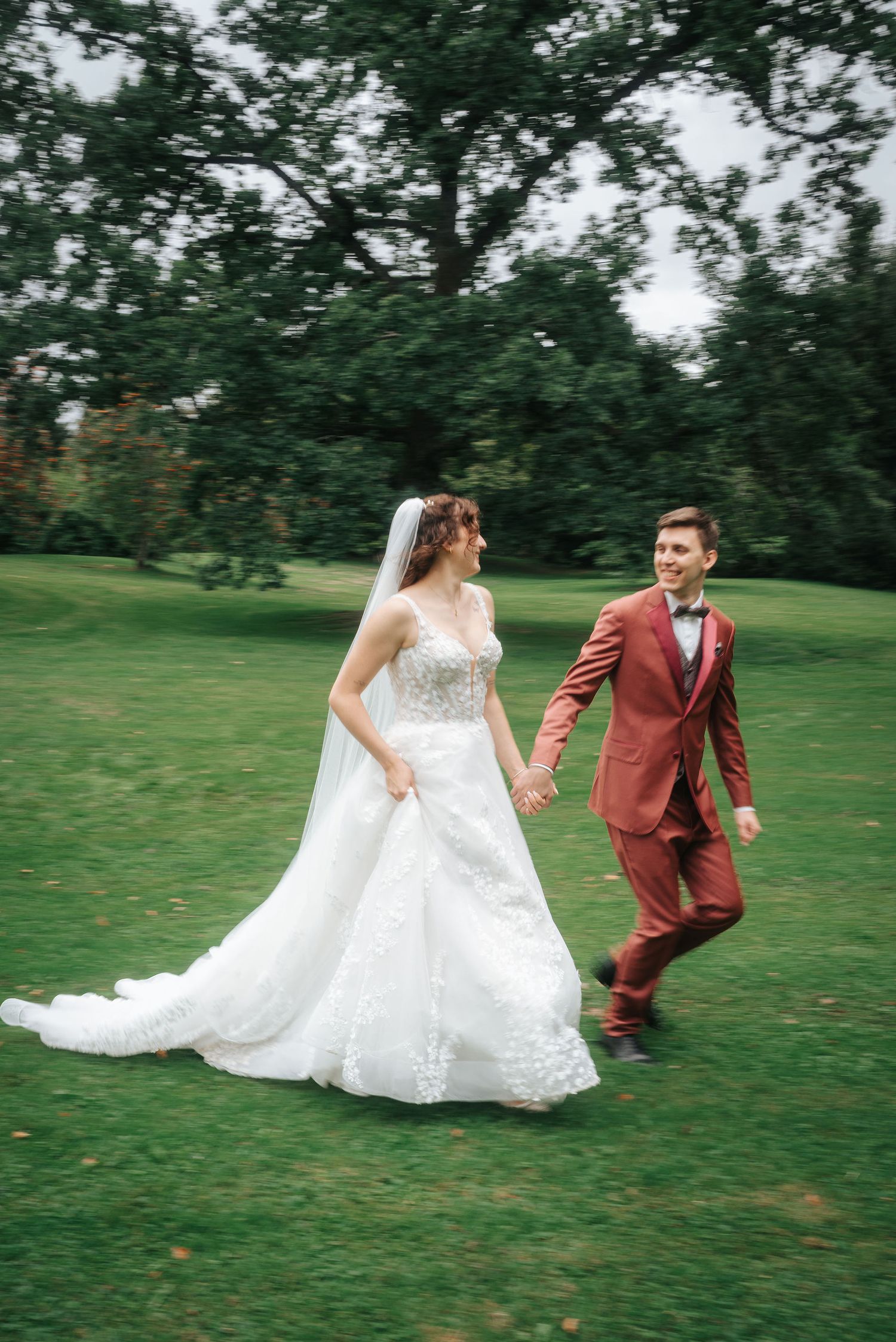 Bride in a lace wedding gown and veil holds hands with a groom in a rust suit as they walk across a green lawn beneath tall trees.