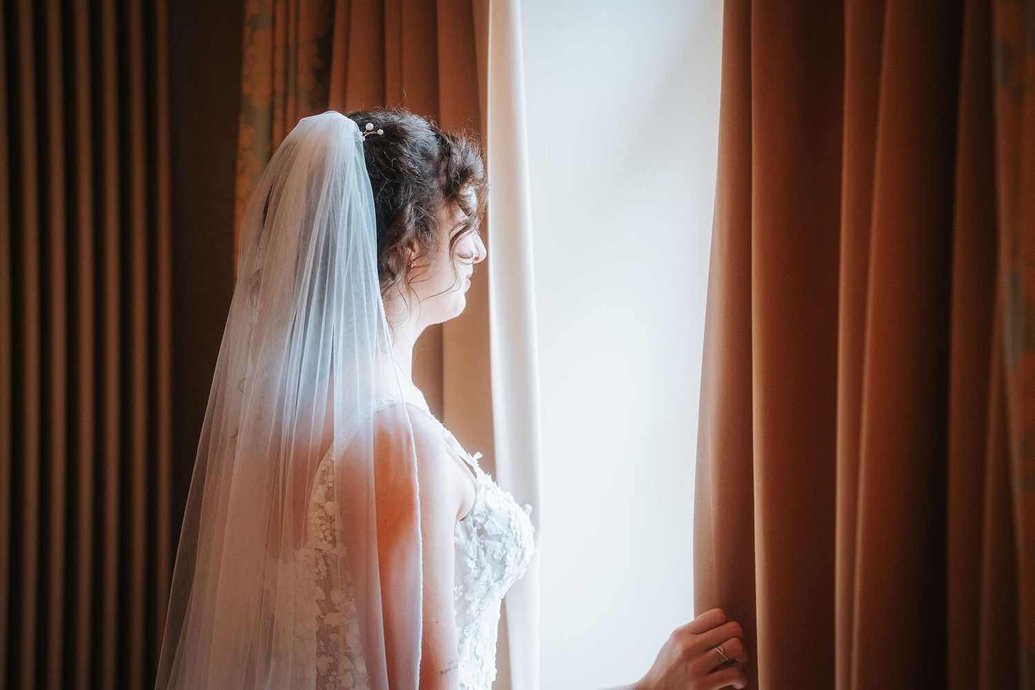 Bride in a lace wedding dress and long veil gazes out a sunlit window beside warm brown curtains.