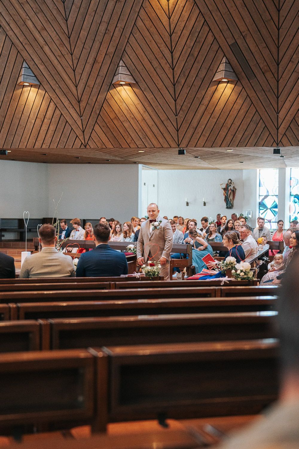 Modern church interior design with wooden herringbone pattern ceiling and pews filled with attendees during a service.