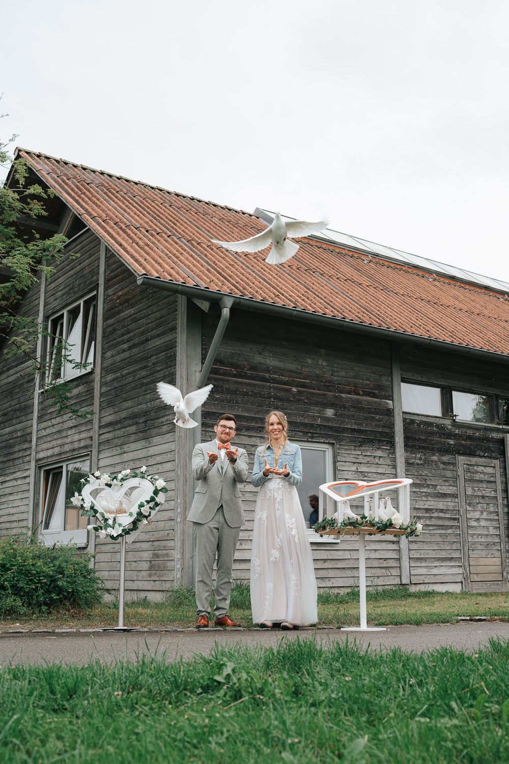Wedding party outdoors posing in front of a rustic wooden barn with terracotta roof tiles and white doves flying overhead.