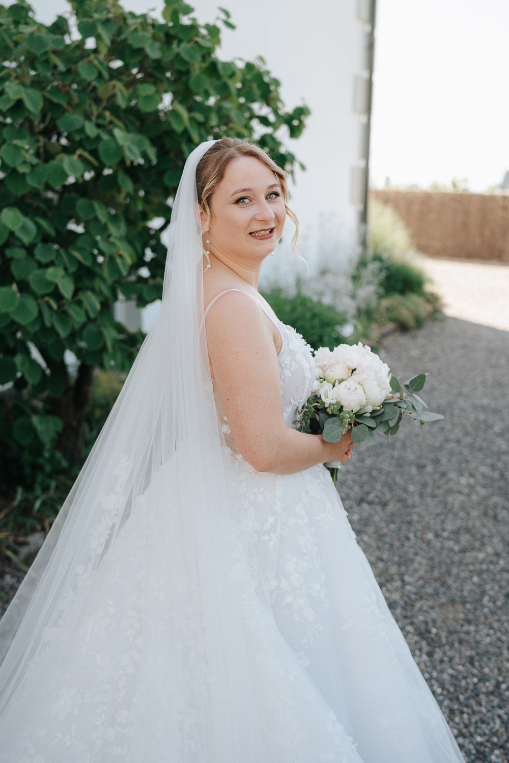 A bride in a classic white ball gown poses outdoors against lush green foliage while holding an elegant bouquet of white flowers.