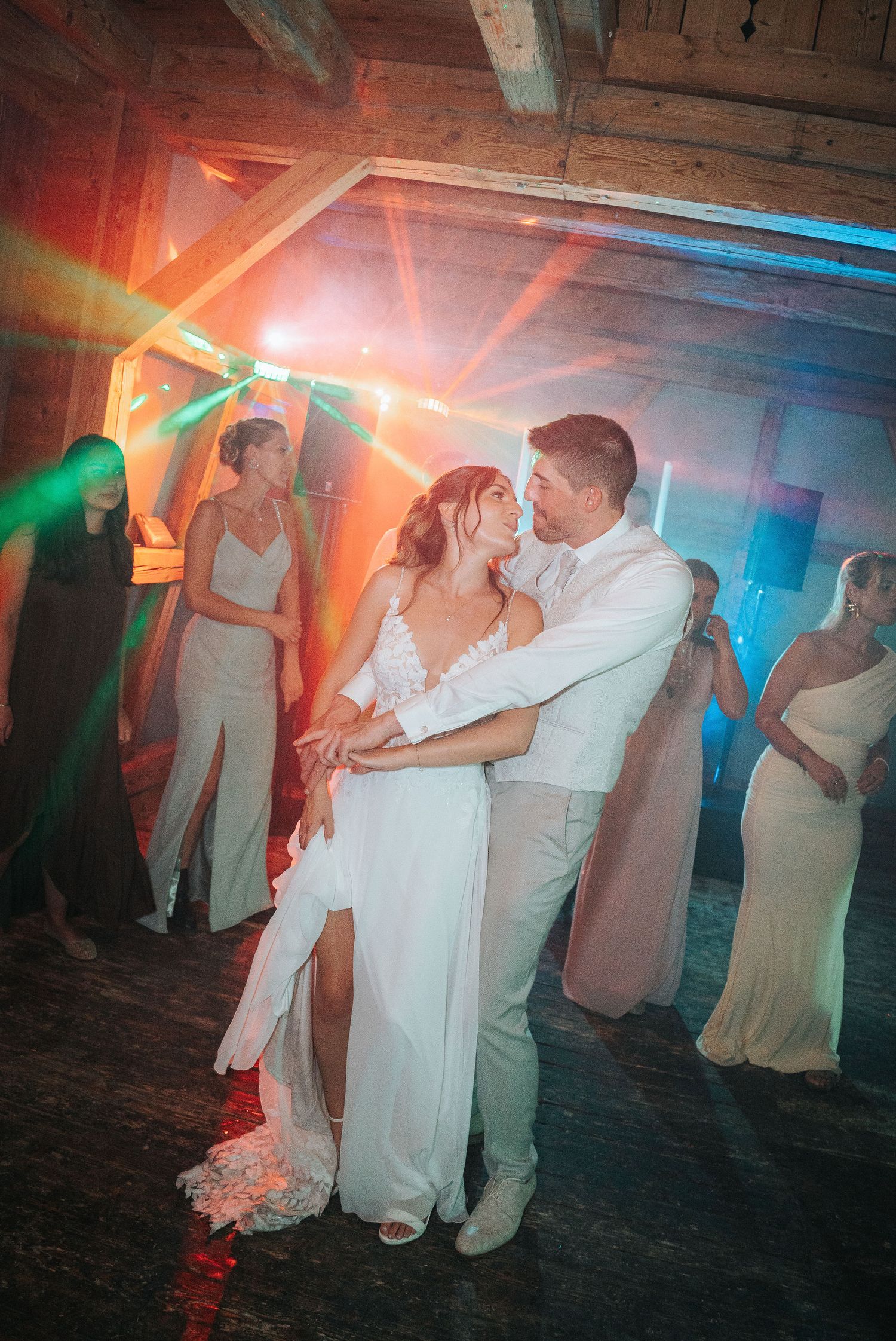 Bride and groom share a romantic first dance in a rustic wooden barn as colorful lights glow and bridesmaids watch.