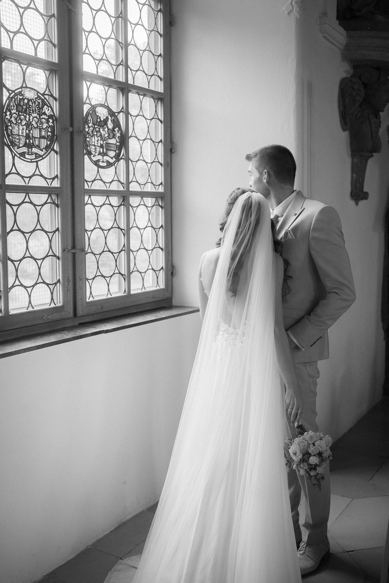 Black and white wedding photo of a groom kissing his bride's forehead beside a leaded glass window, her long veil flowing down.