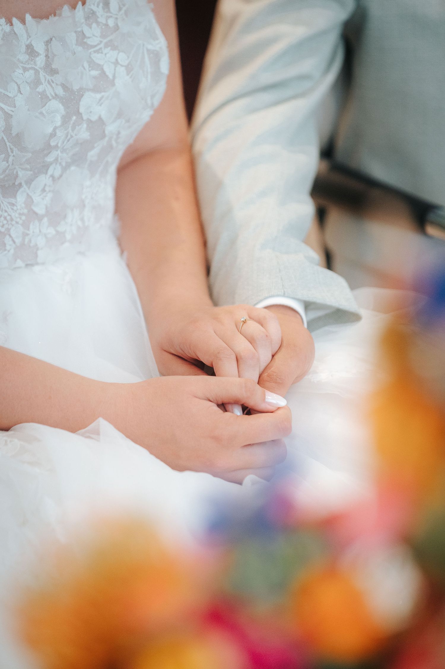 Bride in a white lace wedding dress holds hands with the groom in a gray suit, with colorful flowers blurred in the foreground.