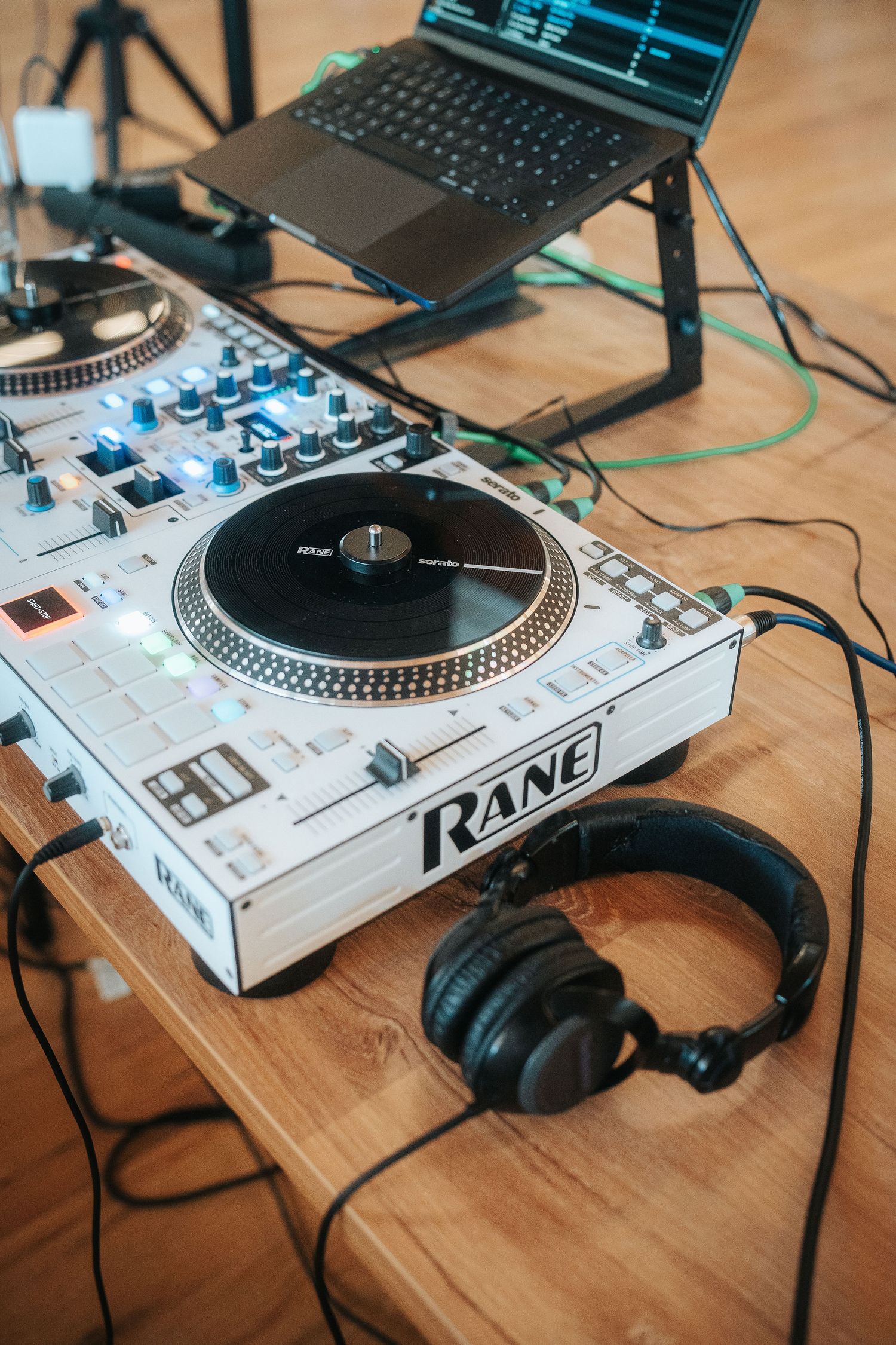 Rane DJ controller with a platter and backlit pads on a wooden desk beside black headphones and a laptop on a stand.