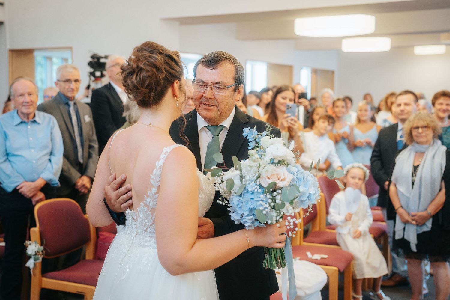 Bride in a white gown holding a blue and white bouquet embraces a smiling man in a dark suit during a wedding ceremony as guests look on.