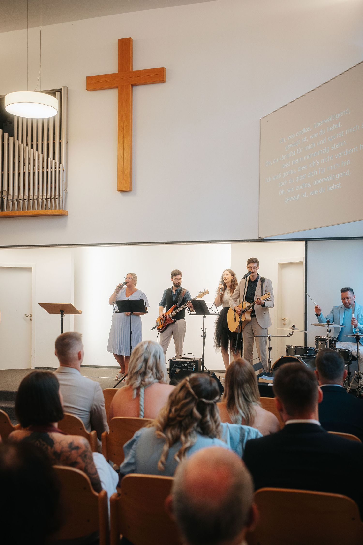 Worship band with singers, guitarists and a drummer performs on stage in a church as the congregation watches beneath a wooden cross.
