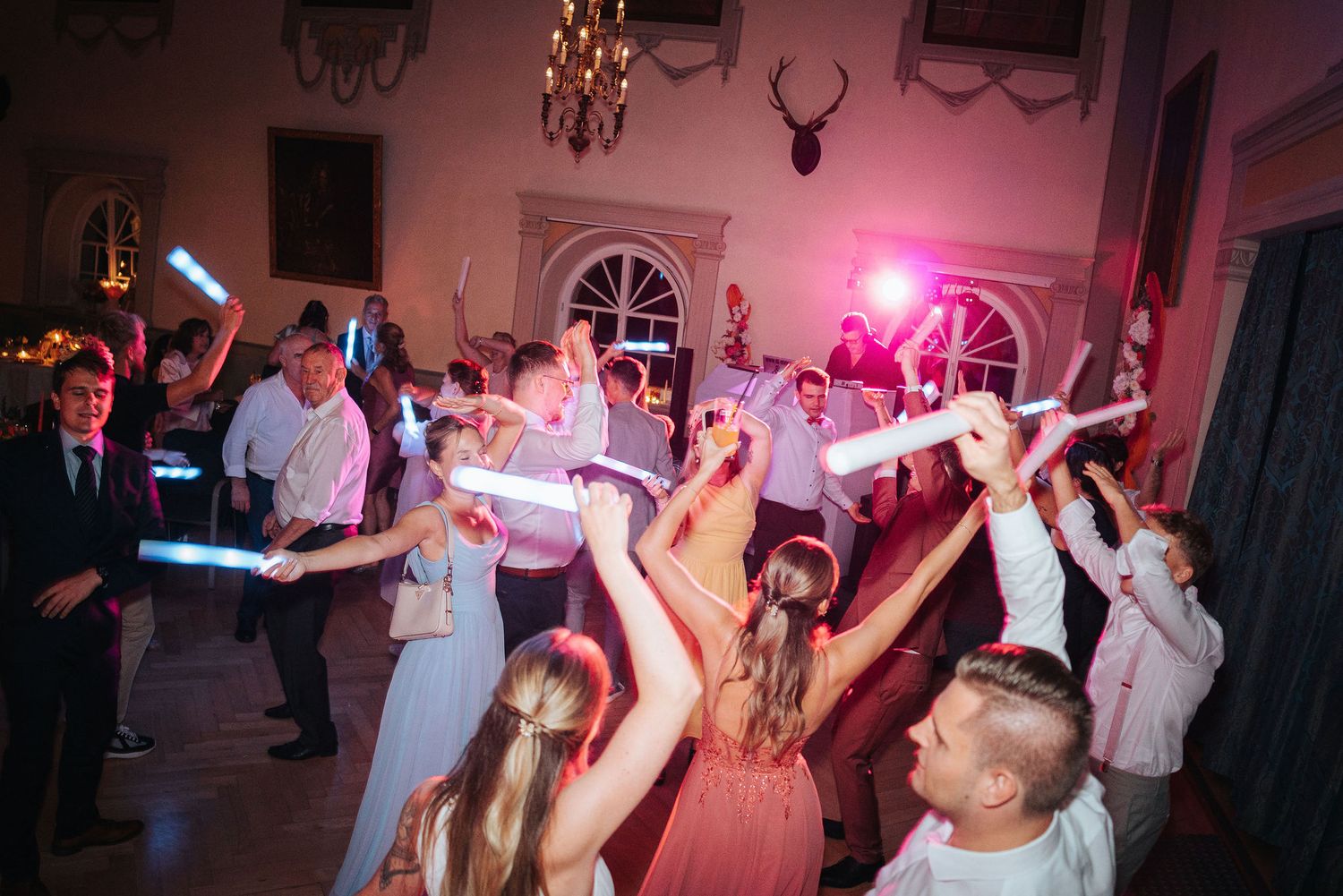 Wedding guests dance with raised arms and glow sticks in a festive, dimly lit ballroom with a mounted antler trophy on the wall.