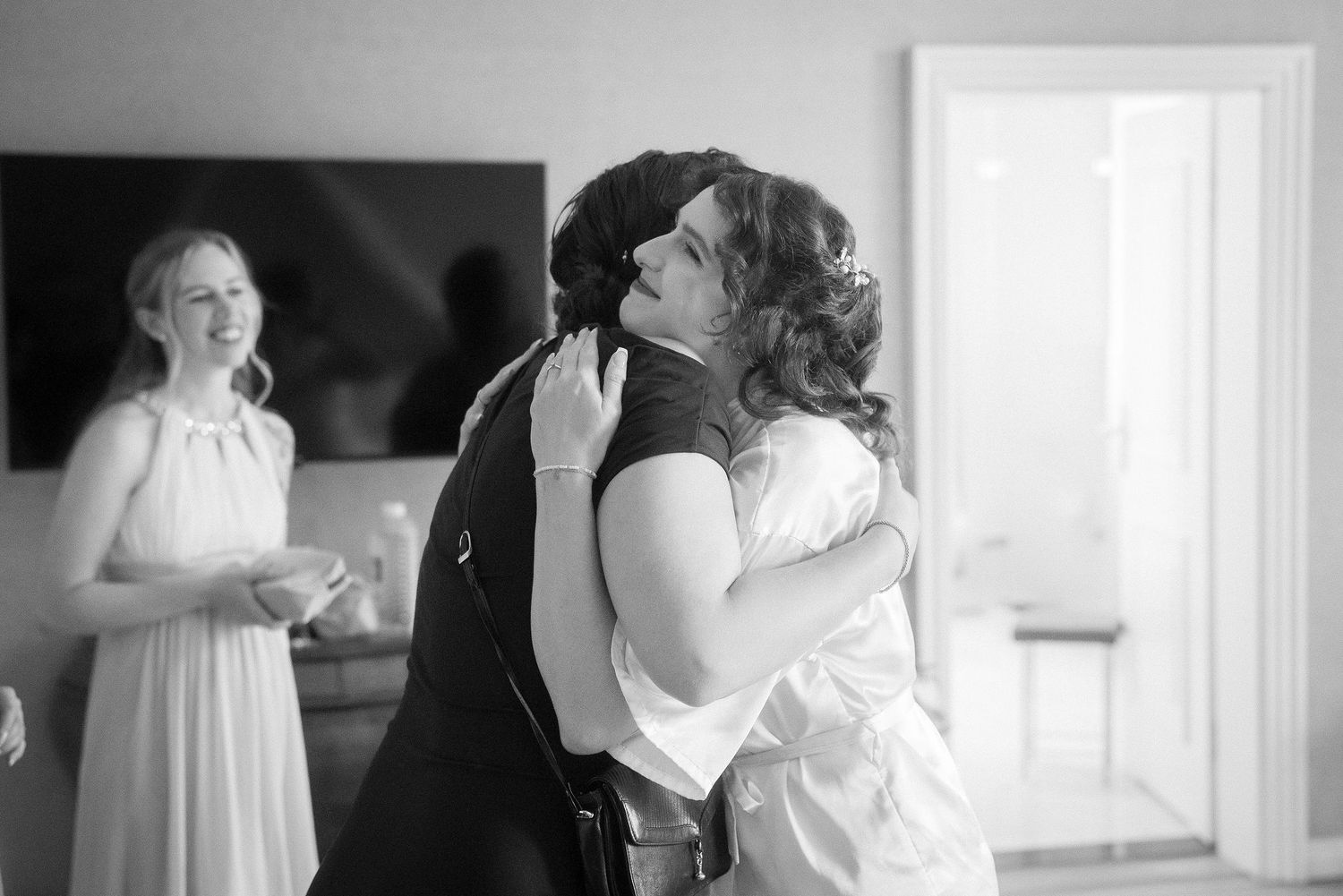 Black and white photo of two women sharing a warm embrace in a bright room while a smiling woman in a long dress watches nearby.