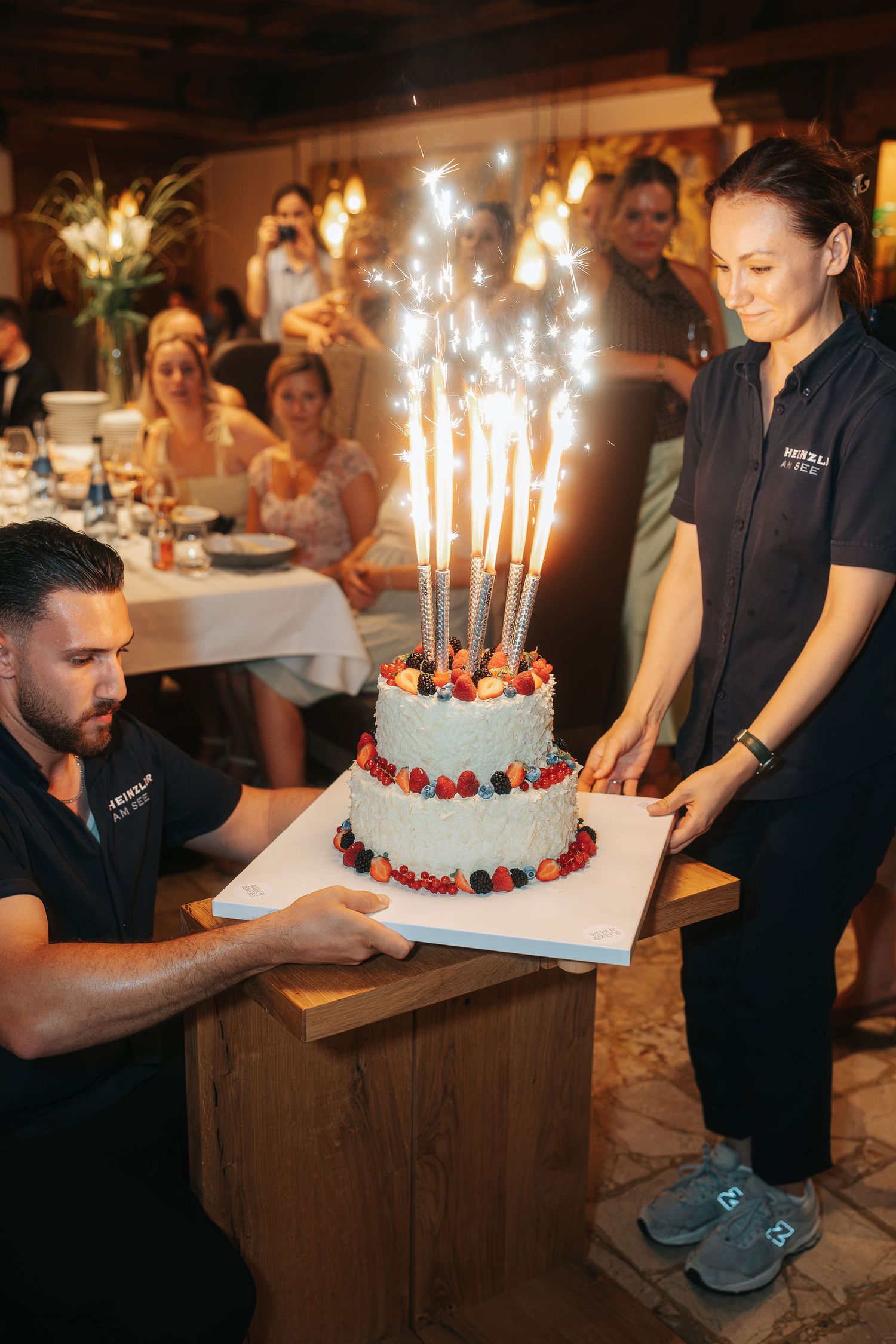 Two staff members in black uniforms present a tiered white celebration cake topped with berries and sparkling candles at a restaurant.