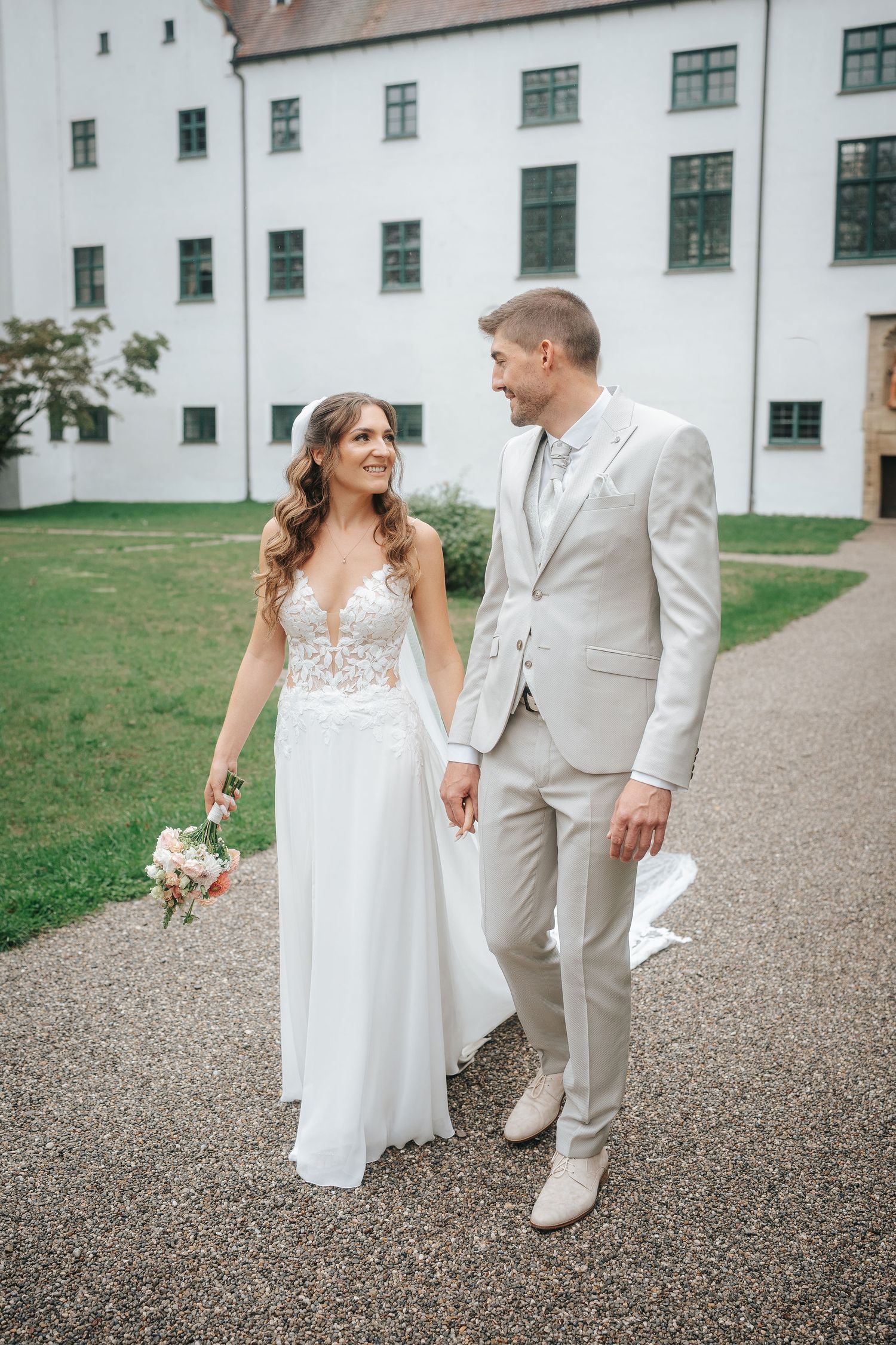 Bride in a lace white gown holding a bouquet walks hand in hand with a groom in a light gray suit past a white castle.