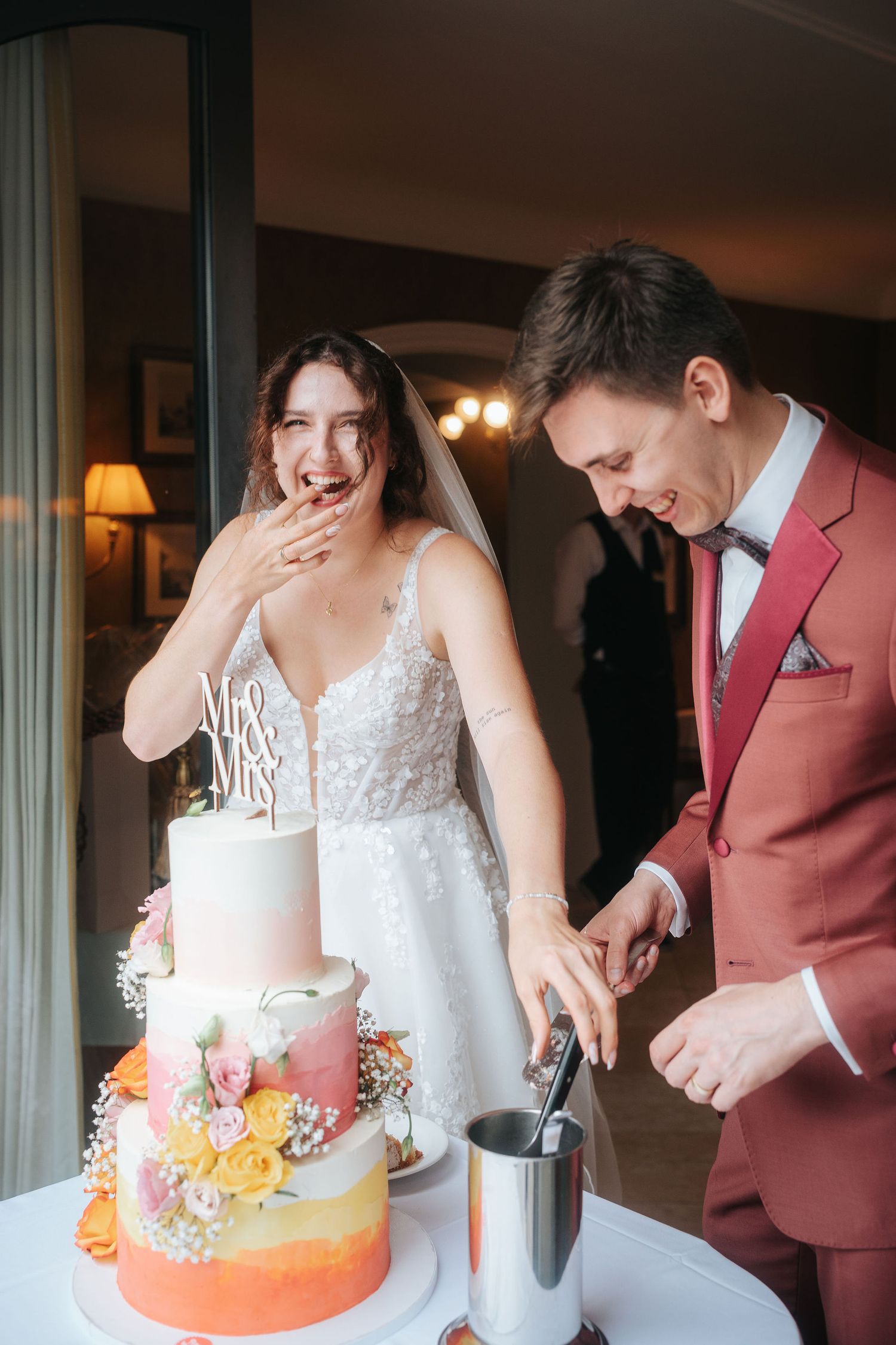 Laughing bride in a lace gown and groom in a rust suit cut their three-tier floral wedding cake topped with a Mr & Mrs sign.
