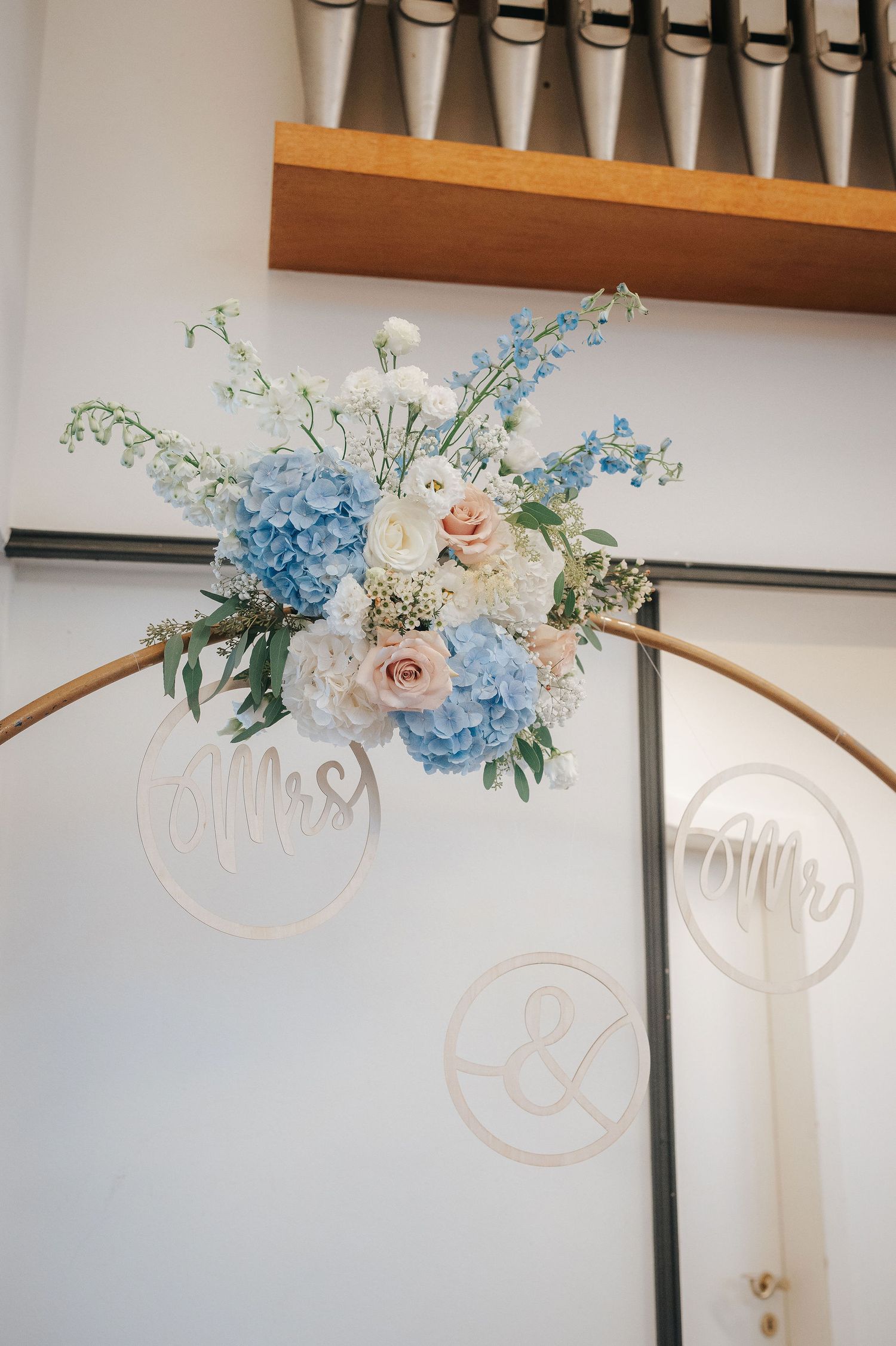 Wedding arch with blue hydrangea, pink roses and white flowers, plus Mr, Mrs and ampersand wooden signs, beneath organ pipes.