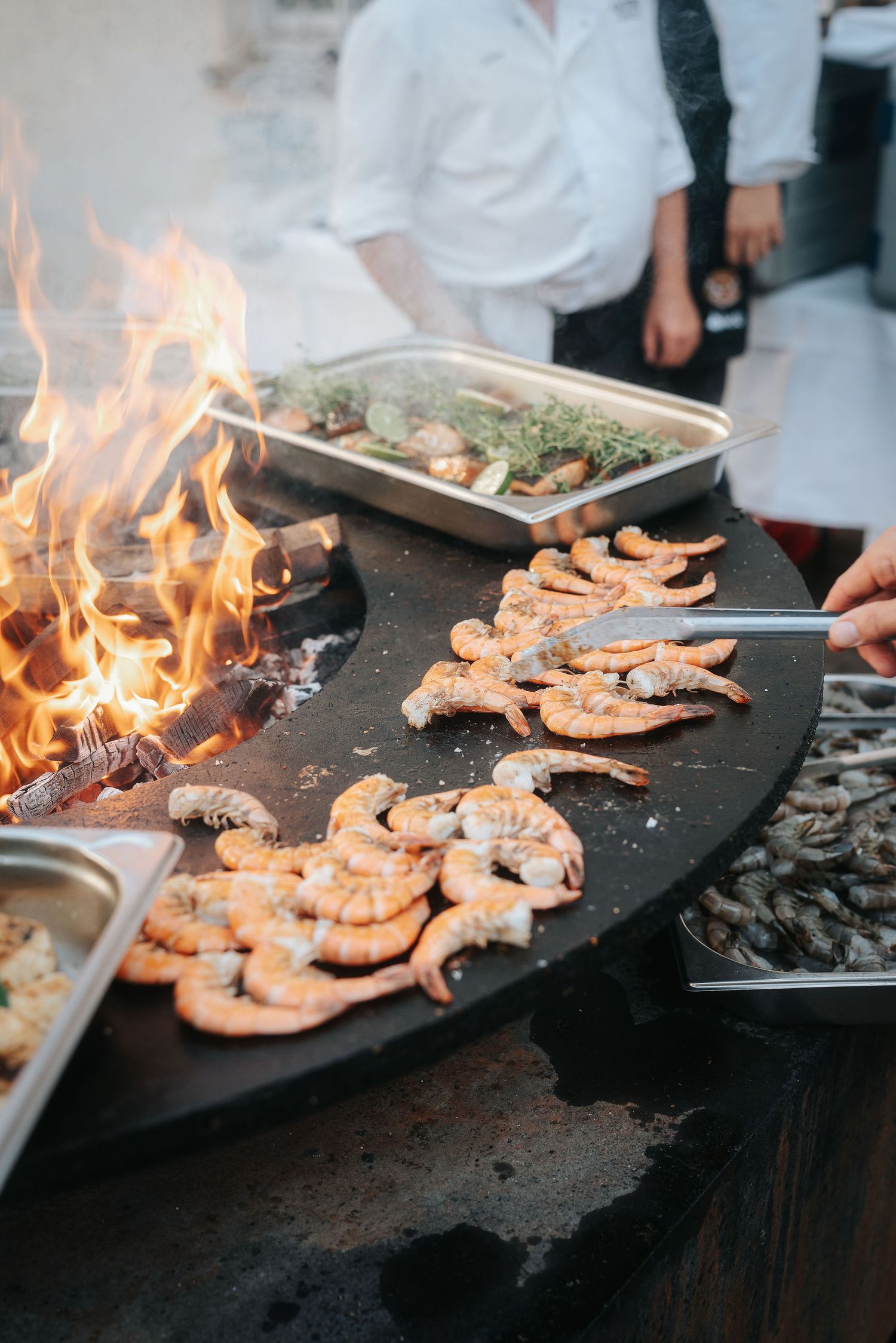 Chefs grill fresh shrimp on a large round flat-top griddle beside an open wood fire, with herb-topped seafood trays nearby.