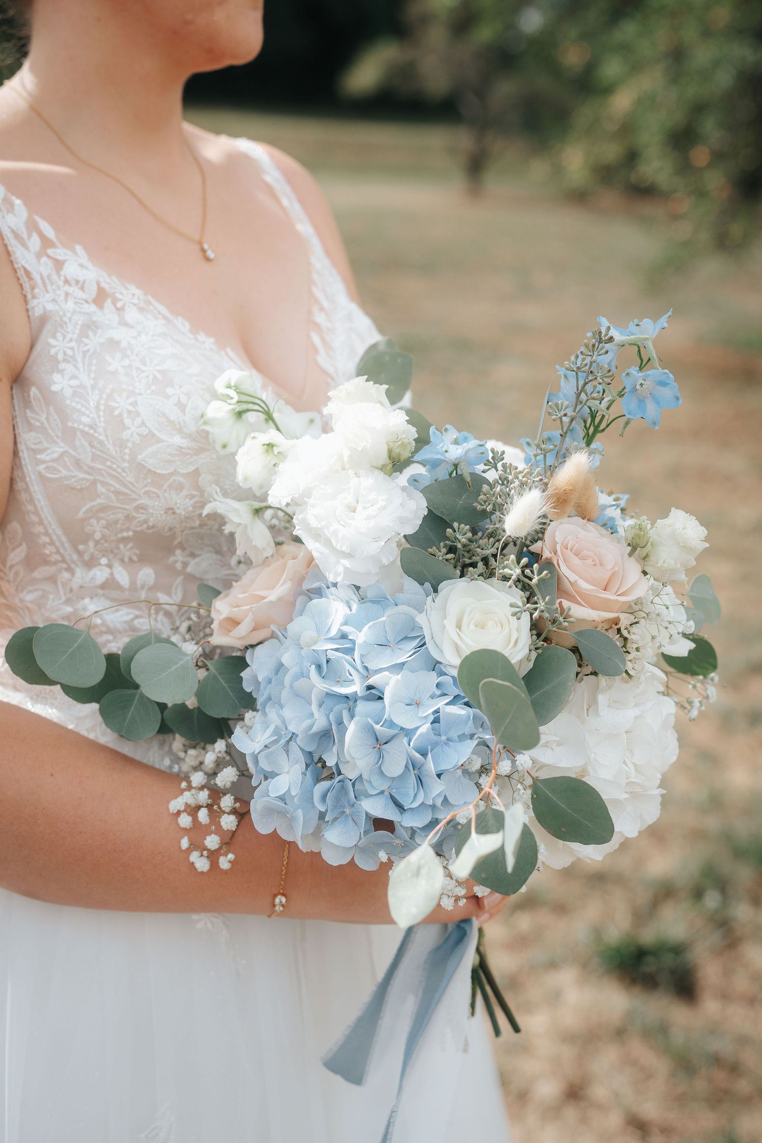 Bride in a lace wedding gown holds a bouquet of blue hydrangea, white roses, blush peonies and eucalyptus tied with a pale blue ribbon.