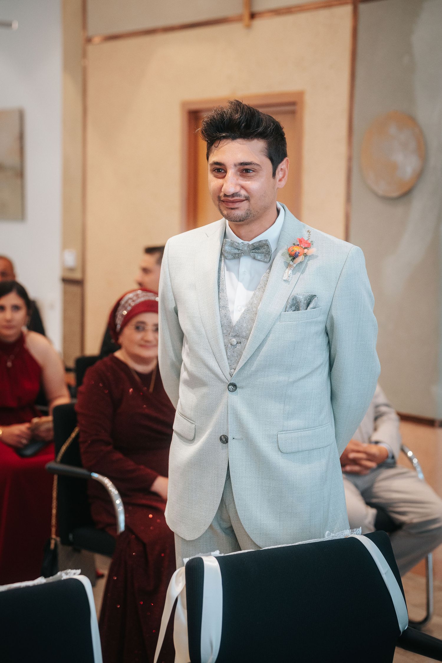 Groom in a light gray suit with a bow tie and floral boutonniere stands smiling during a wedding ceremony as guests sit behind him.