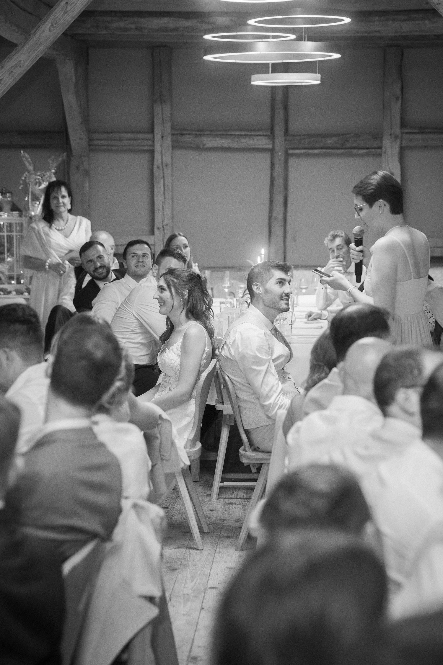 Black and white photo of wedding guests seated at a rustic barn reception as a woman gives a speech under modern ring lights.