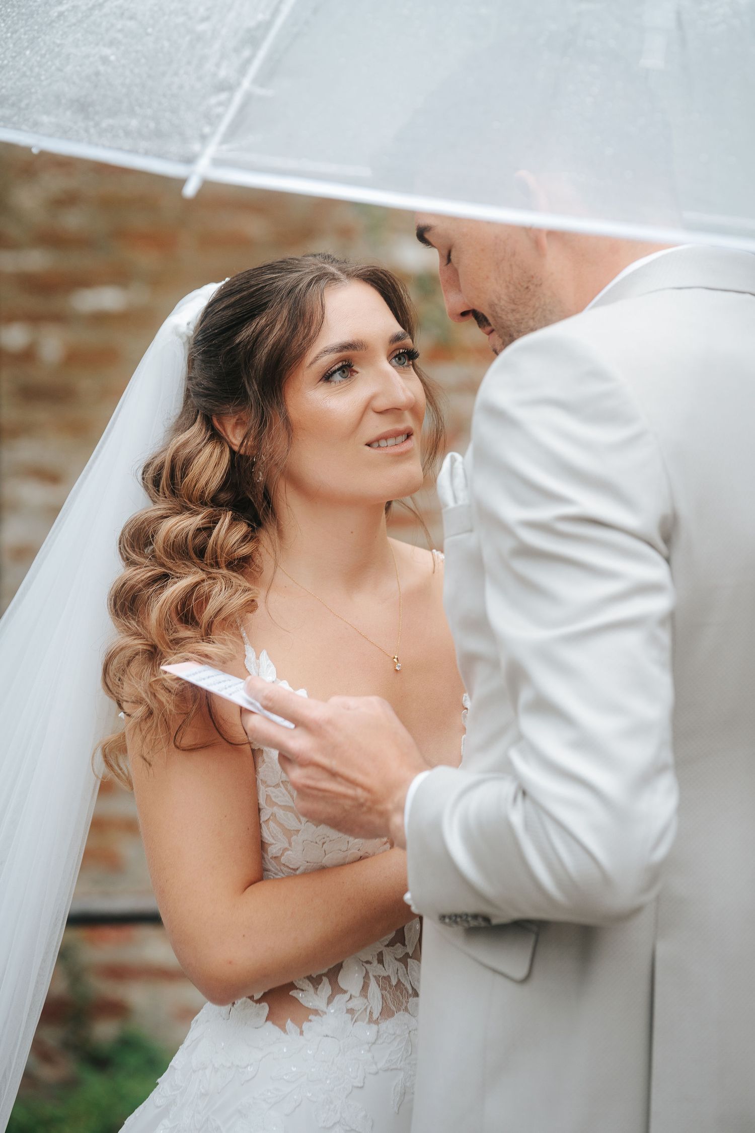 Bride in a lace wedding gown and veil gazes up at a groom in a light suit as they stand together under a clear umbrella.