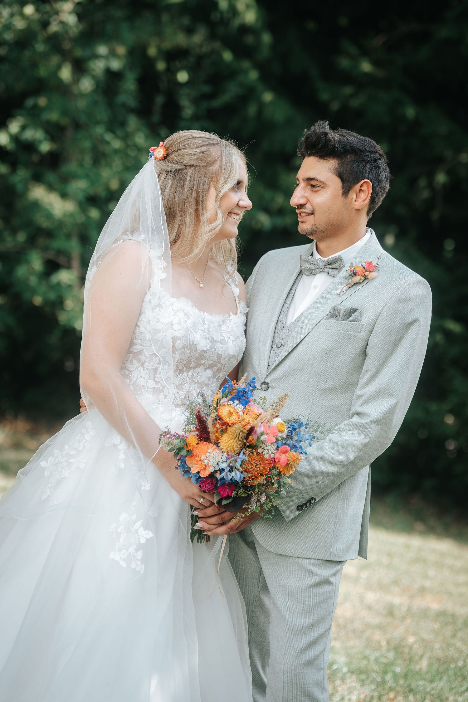 Smiling bride in a lace wedding gown and veil holds a colorful wildflower bouquet while gazing at the groom in a light gray suit outdoors.