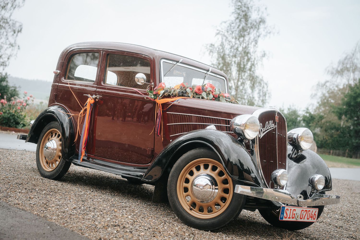 Vintage burgundy wedding car with floral decoration and orange ribbons parked on a gravel driveway surrounded by trees.
