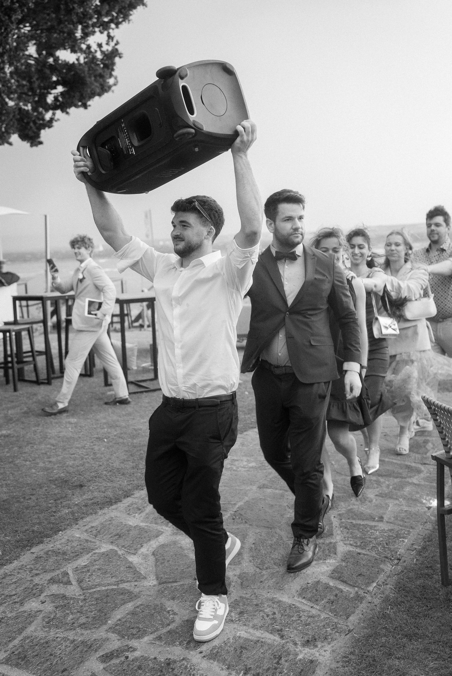 Black and white photo of a man in a white shirt carrying a large speaker overhead as wedding guests follow him in a lively procession.