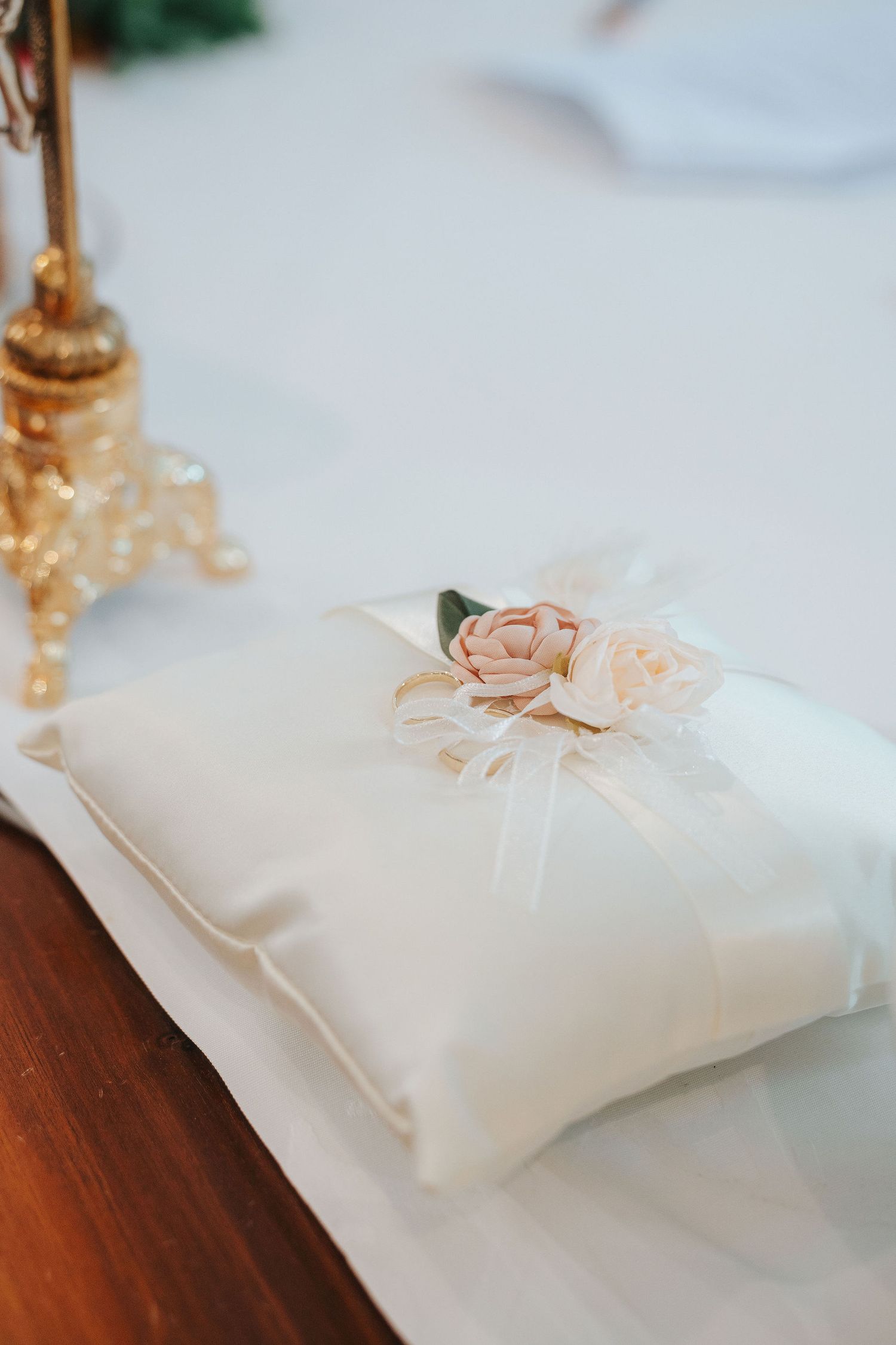 Cream satin wedding ring bearer pillow adorned with soft pink and ivory fabric roses and sheer ribbon, resting on a table beside gold decor.