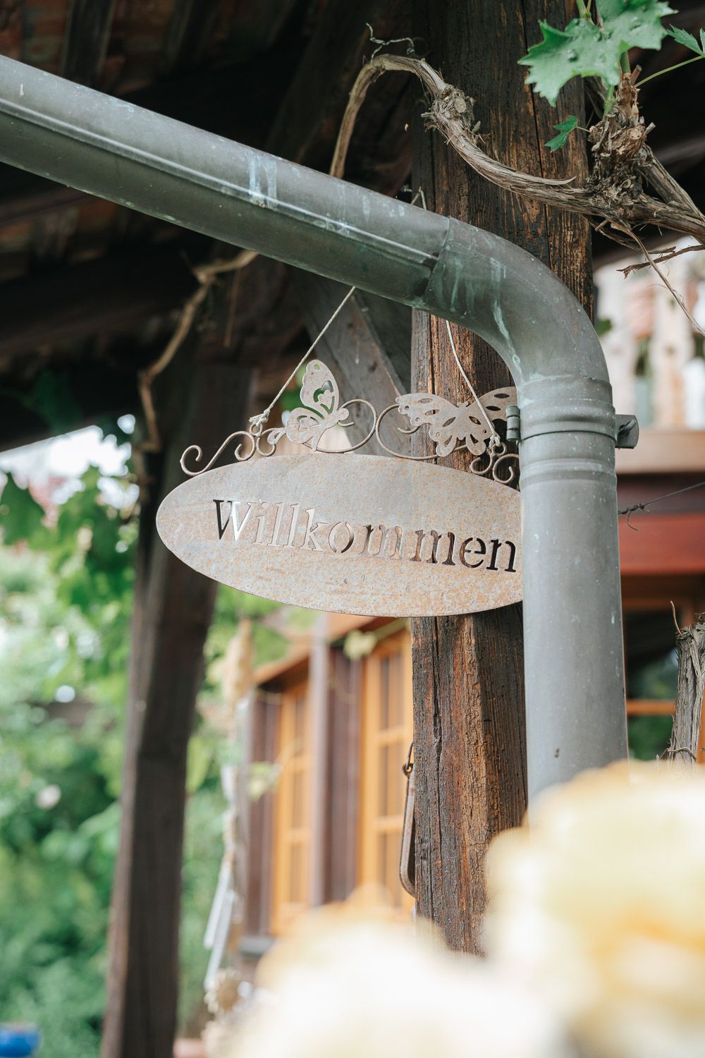A decorative metal welcome sign with the text 'Willkommen' hangs from a rustic wooden beam outdoors.