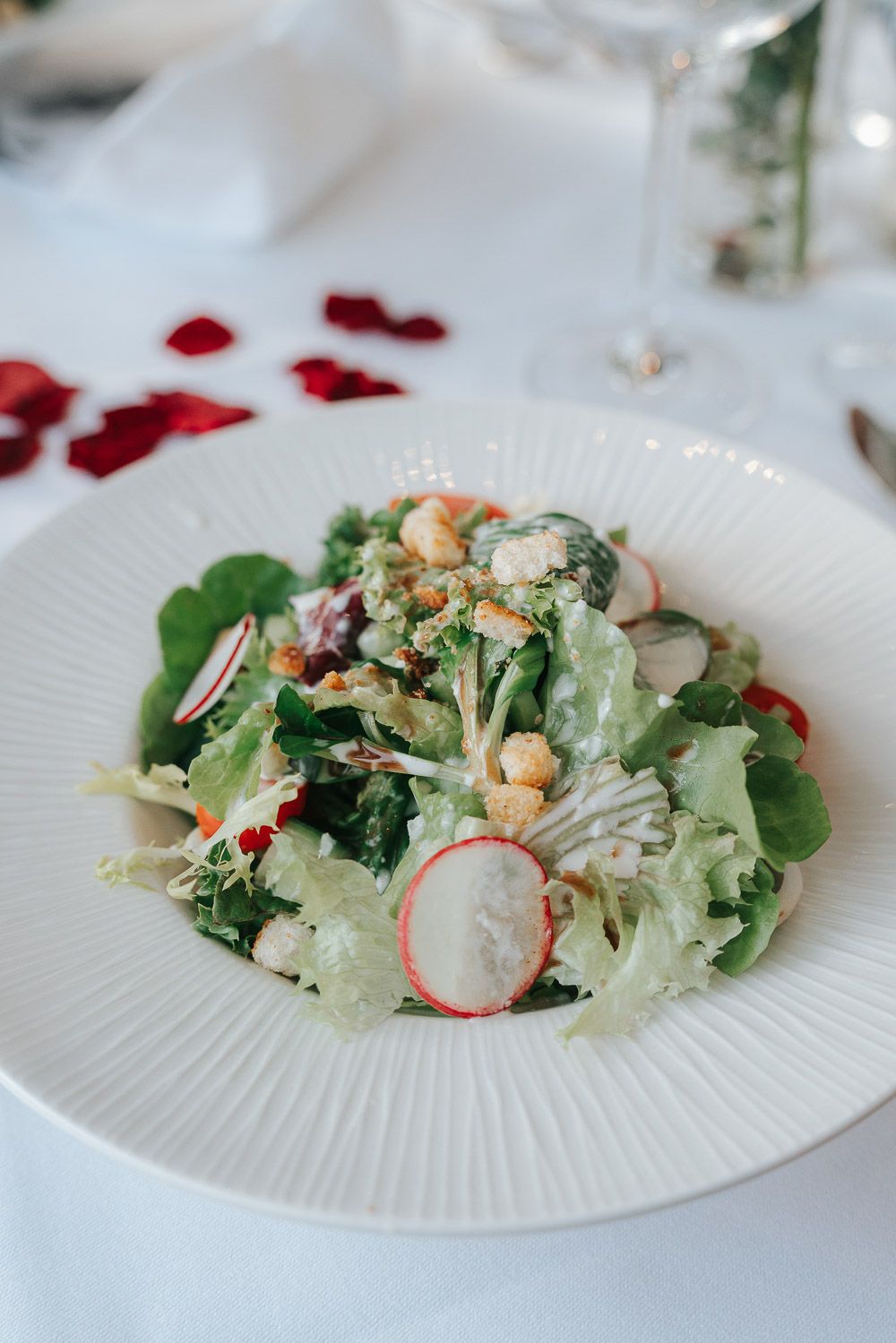 Fresh garden salad with sliced radishes, greens, croutons and nuts served on a white ribbed plate with rose petals nearby.