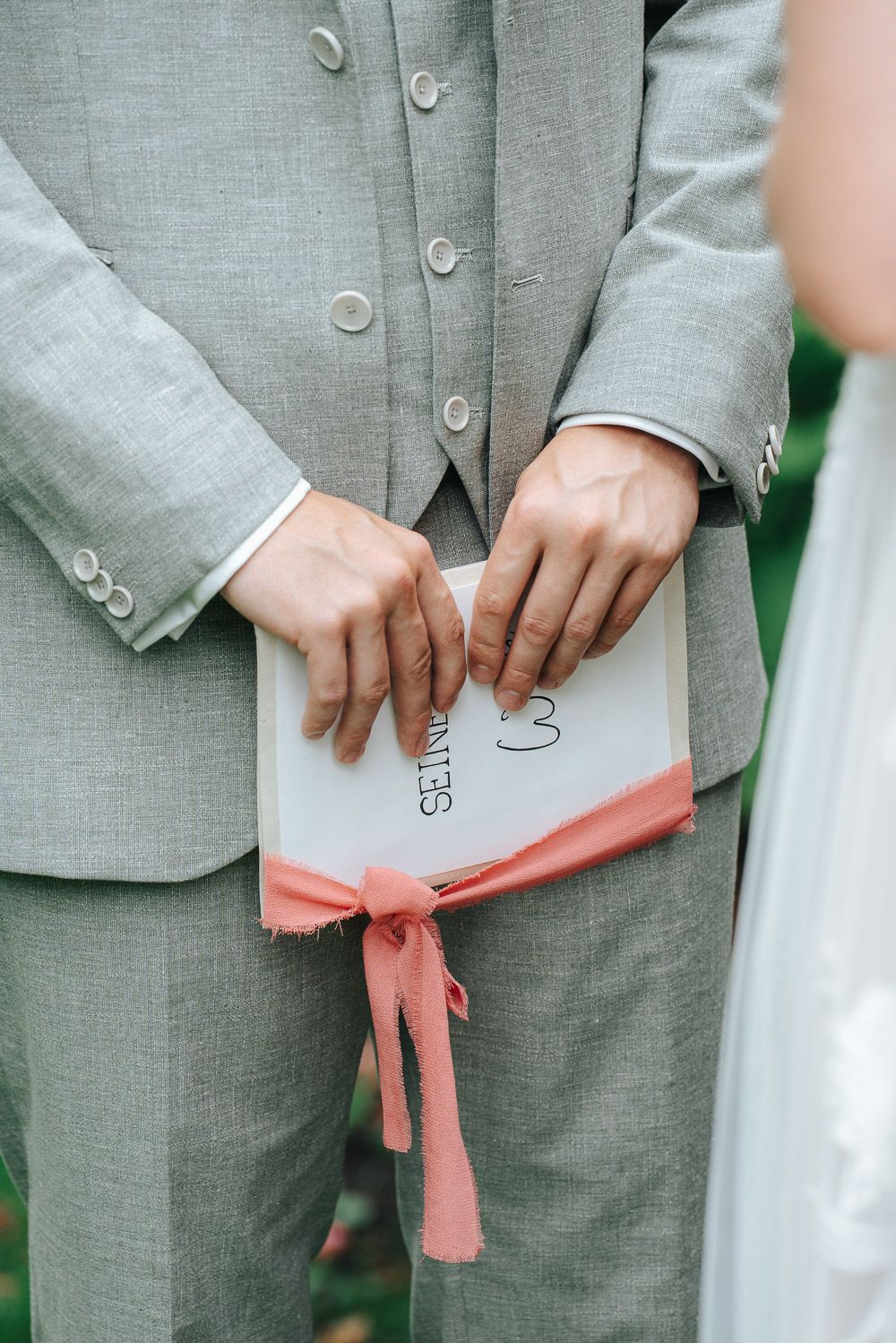 Hands holding a small statement card with a pink ribbon and wearing a light gray suit.