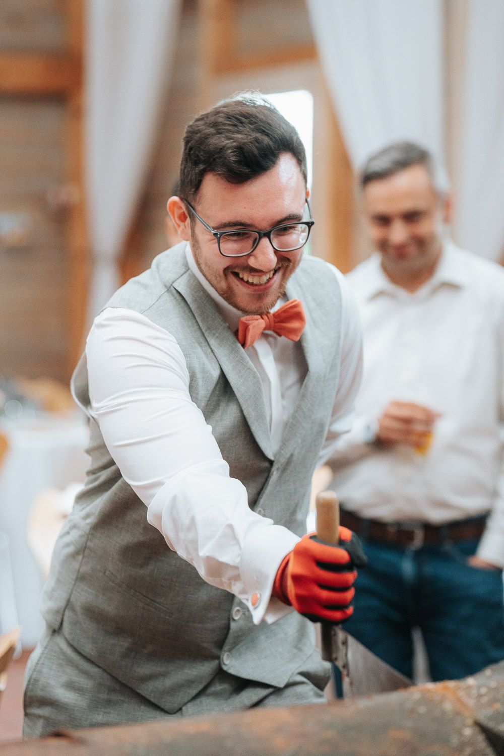 A groom in a grey vest and bow tie wearing red work gloves while chopping wood at a wedding.