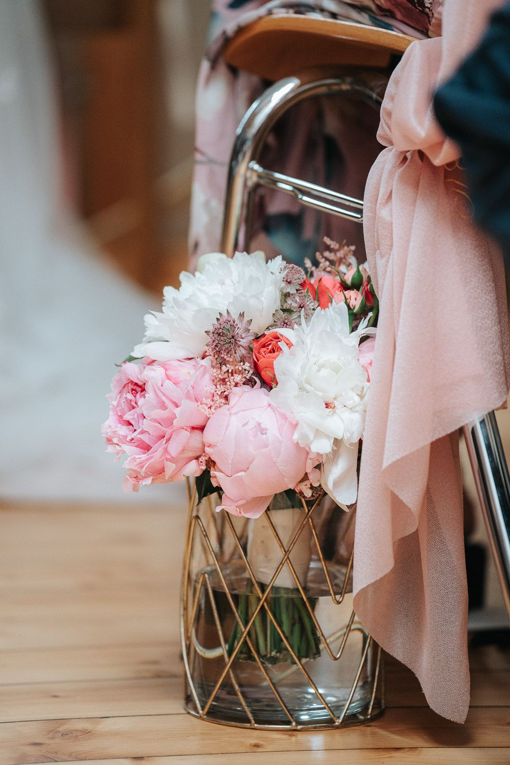 Pink peonies and white flowers in a geometric glass vase with pink fabric draped beside it on wooden surface.