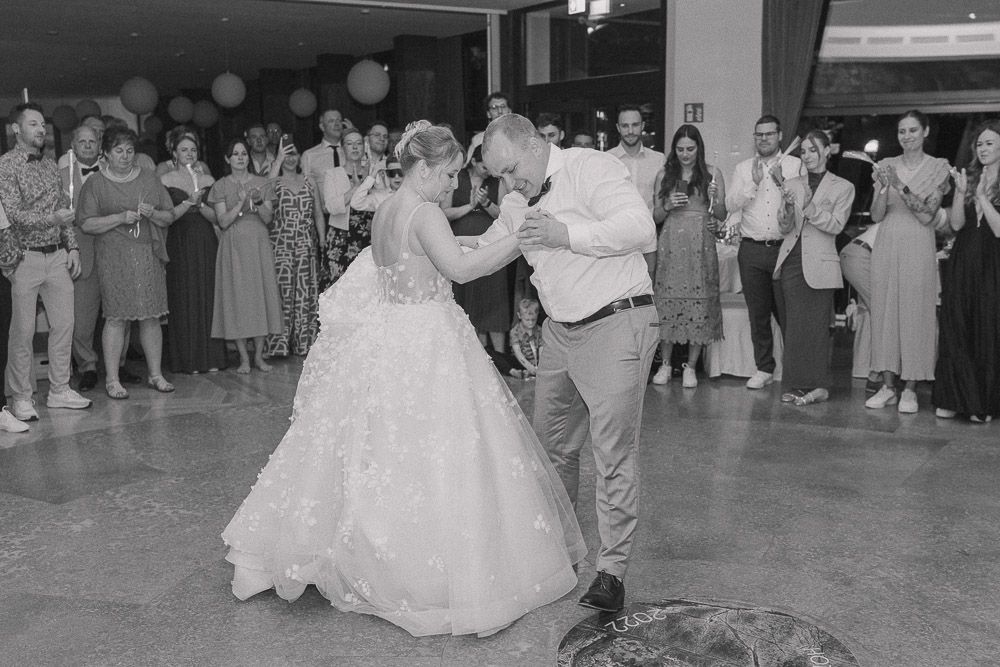A couple dances their first dance at a wedding reception while guests watch and take photos in black and white.