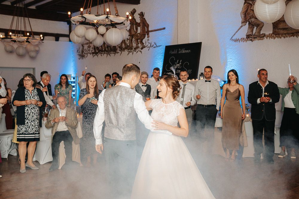 A couple dances their first dance at a wedding reception as guests watch in a misty venue with ornate chandeliers.