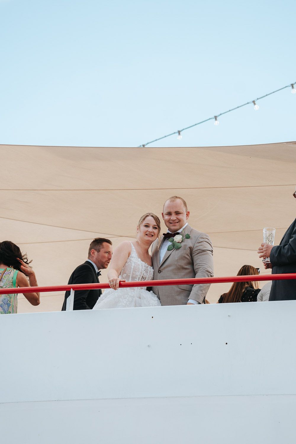 A wedding celebration scene shows people joyfully posing and interacting along a white railing against a beige wall.