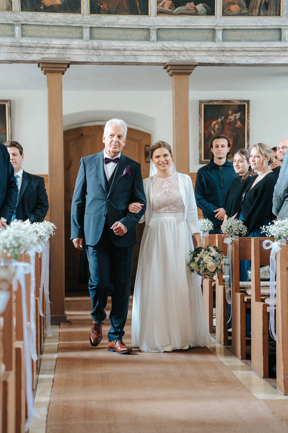 A collage of five wedding processional photos showing ceremony guests walking down a church aisle decorated with flowers.