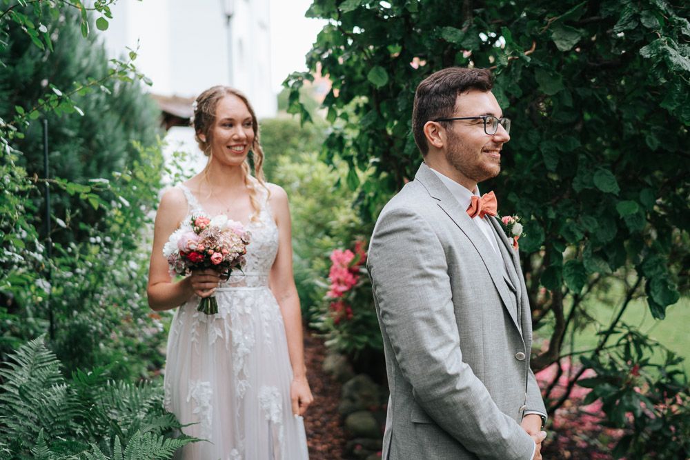 Wedding couple stands together in a lush garden setting under dappled sunlight in elegant white and gray attire.