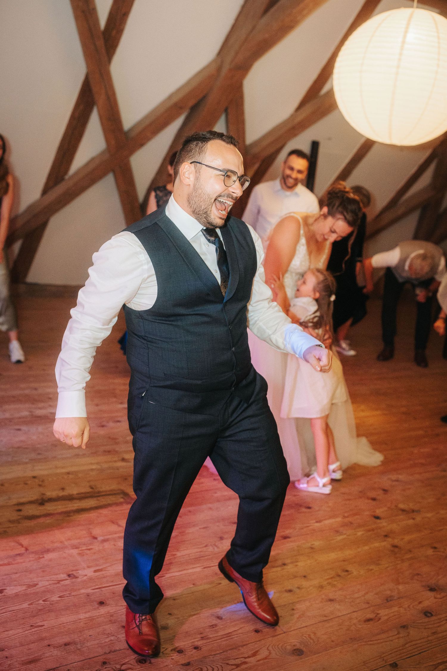 Smiling bearded man in a navy vest and brown shoes dances joyfully at a wedding reception in a rustic wooden-beamed venue.