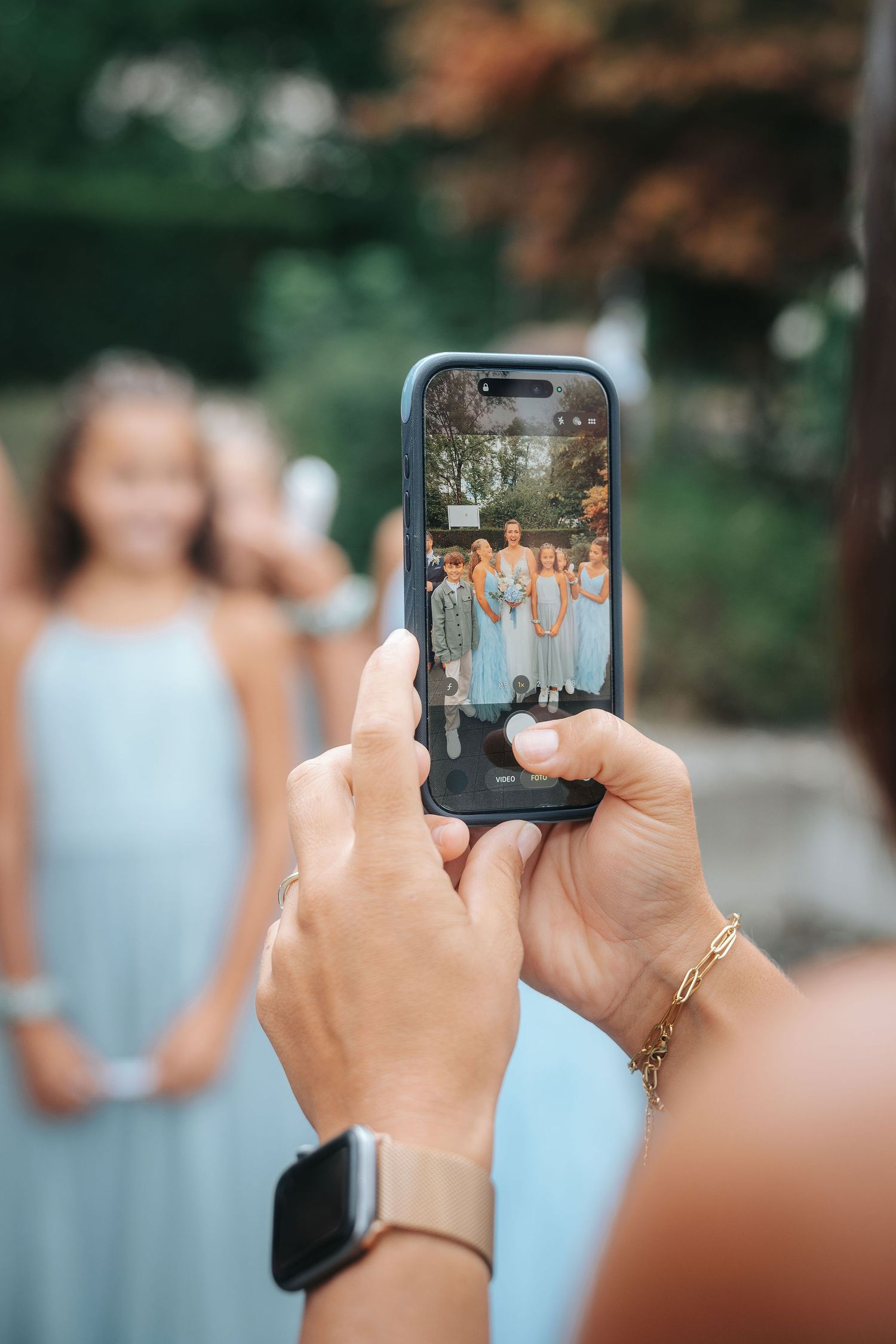 Woman photographs bridesmaids in light blue dresses on her smartphone at an outdoor wedding, with the phone screen showing the group.