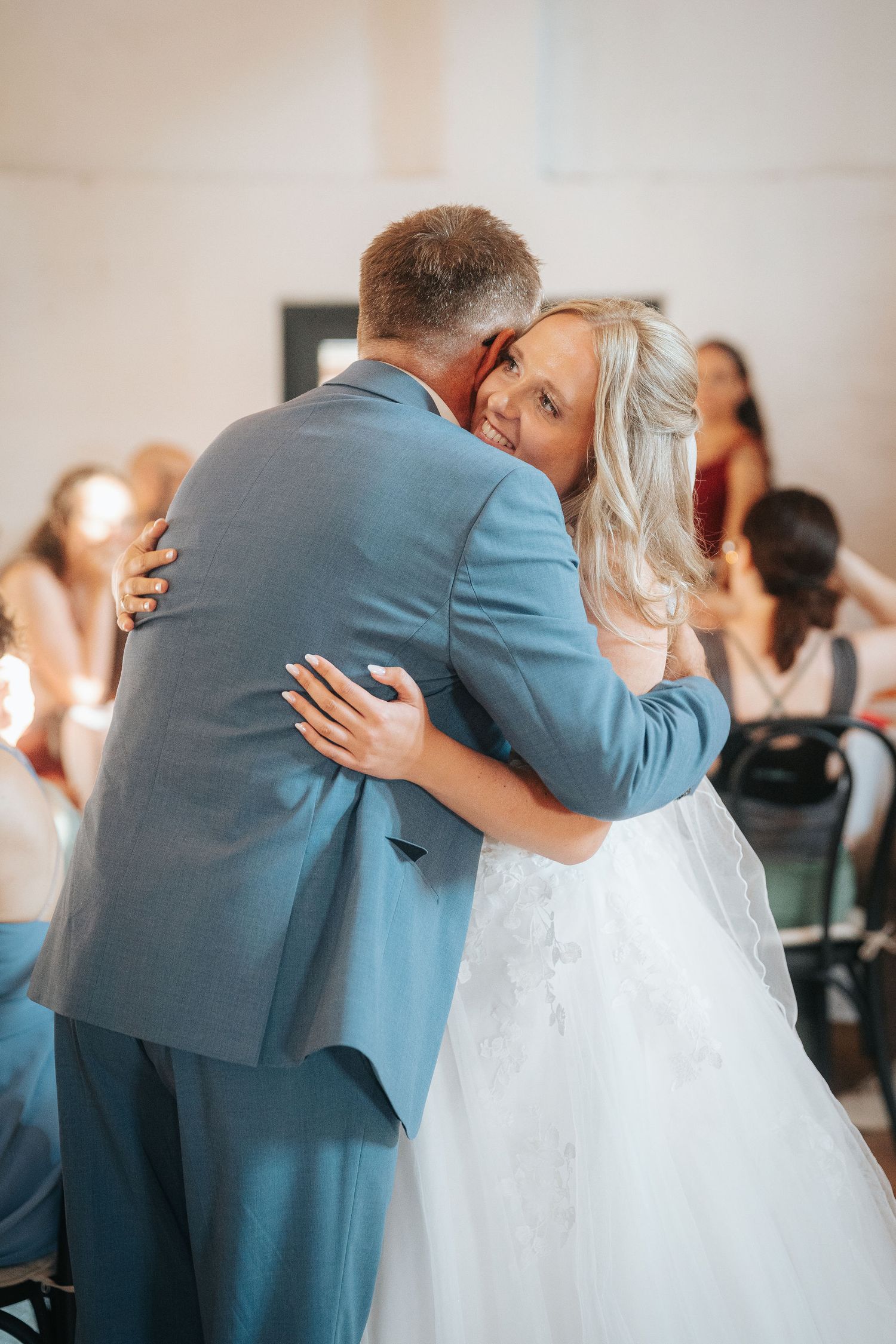 Smiling bride in a white lace wedding gown shares a warm embrace with a man in a blue suit during a reception dance as guests look on.