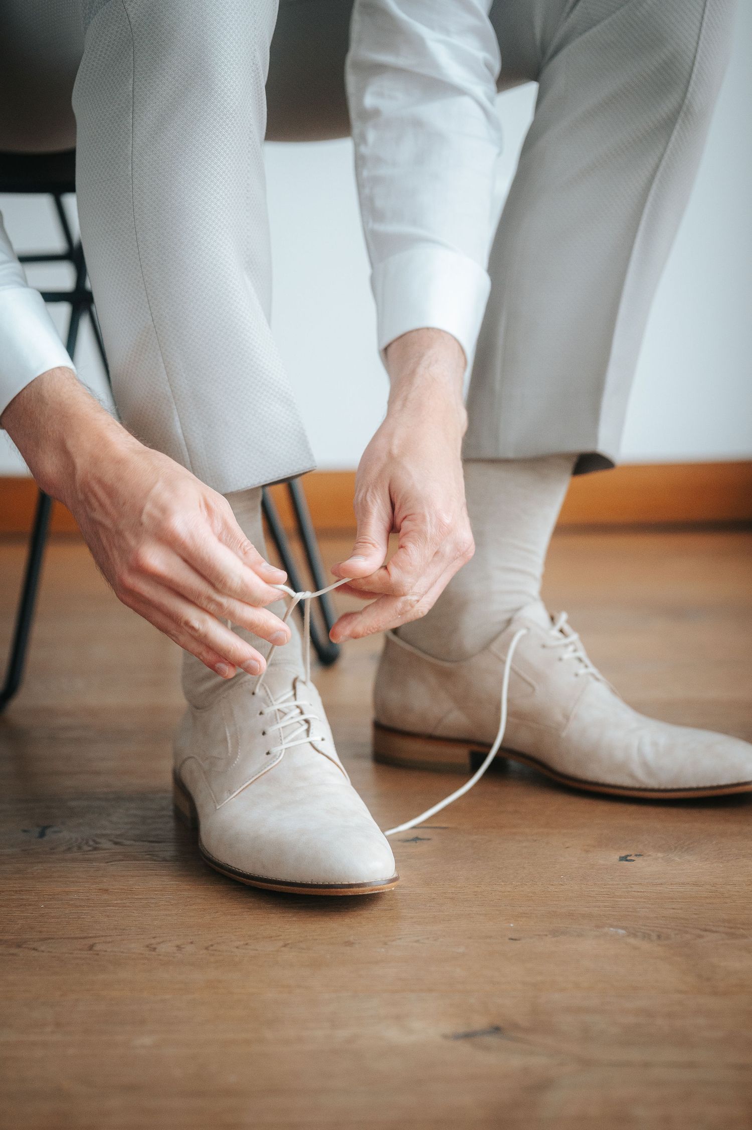 Man in light gray trousers and a white shirt sits on a chair, tying the laces of beige suede dress shoes on a wooden floor.