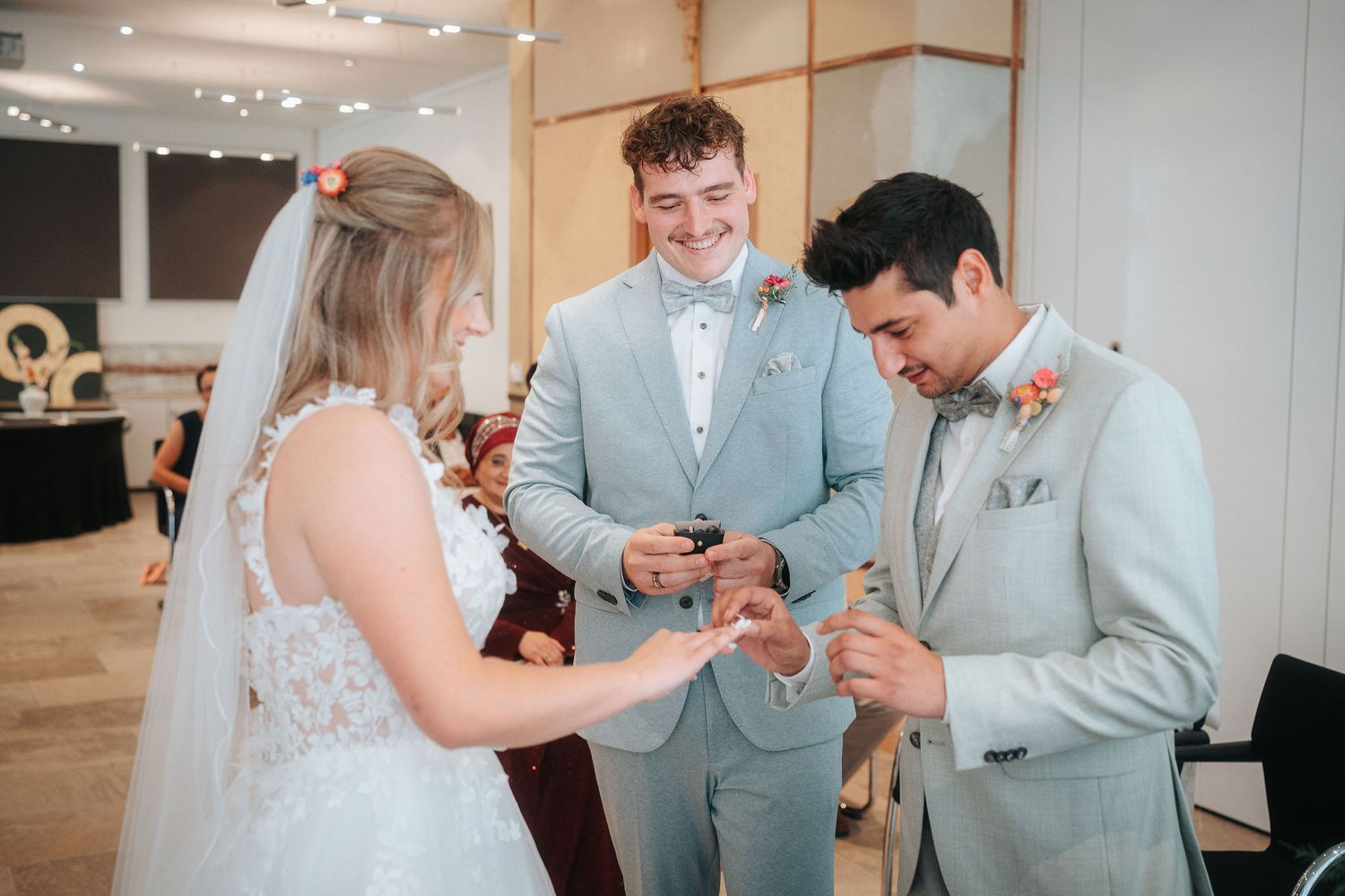 Bride in a white lace gown and veil exchanges rings with a groom in a gray suit while a smiling groomsman looks on.