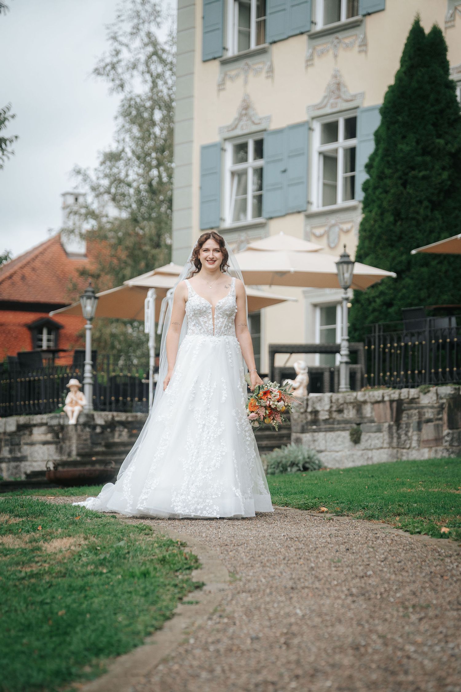 Smiling bride in a white lace wedding gown holding a colorful bouquet on a gravel path outside a historic building with blue shutters.