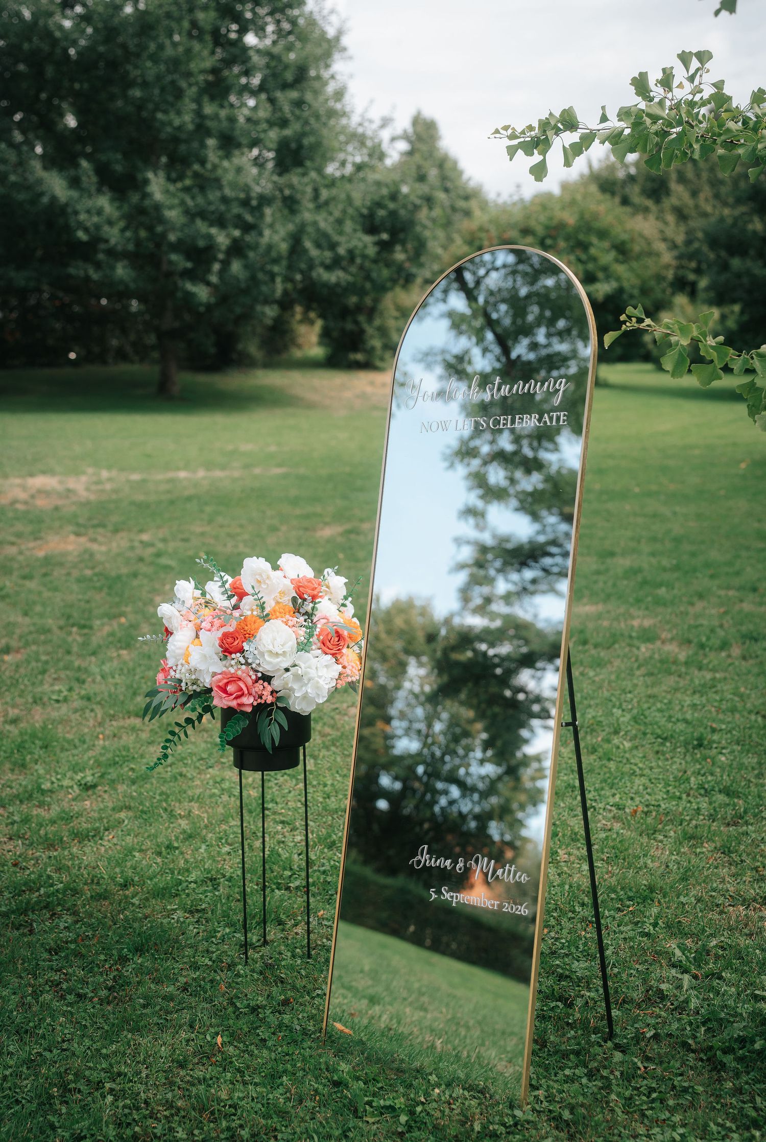 Arched gold-framed wedding welcome mirror with white script beside a black stand of peach and white flowers on a sunny lawn.