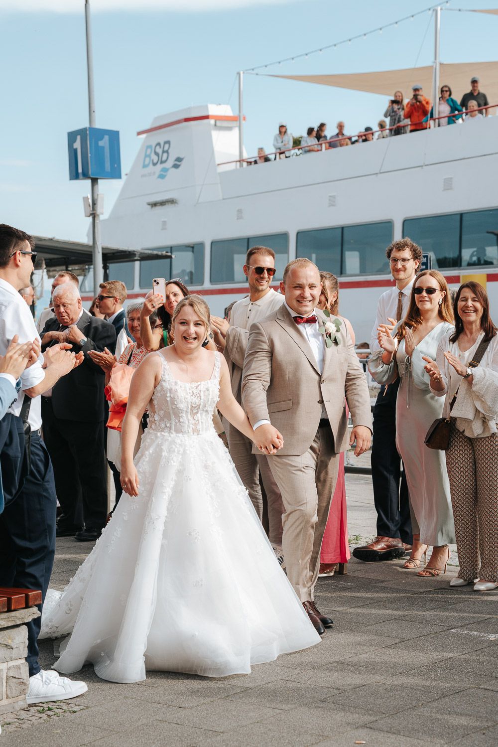 A wedding celebration takes place outside with guests looking on as a couple walks together in formal attire.