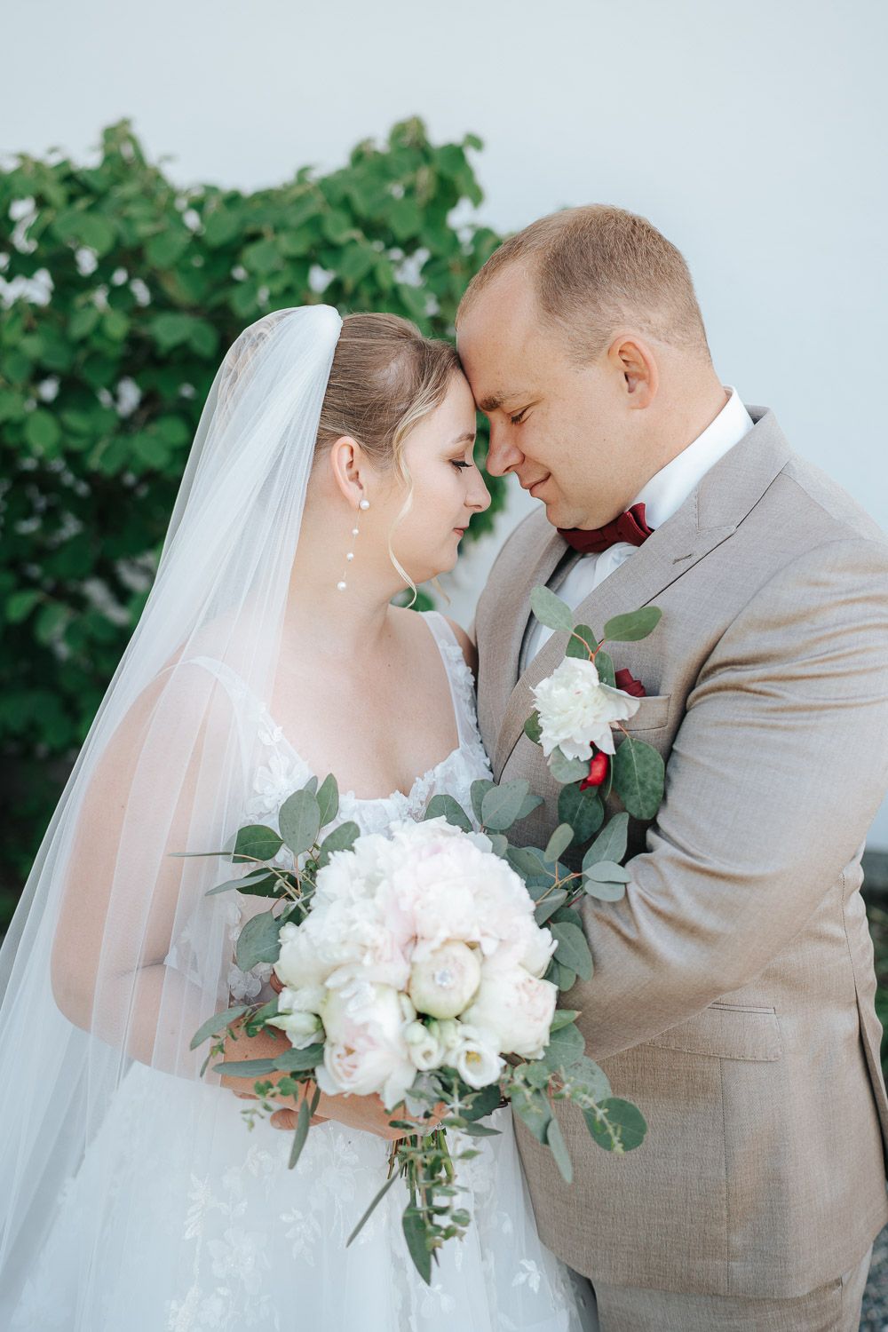 Newlywed couple shares intimate moment in garden setting with eucalyptus and white rose bouquet.