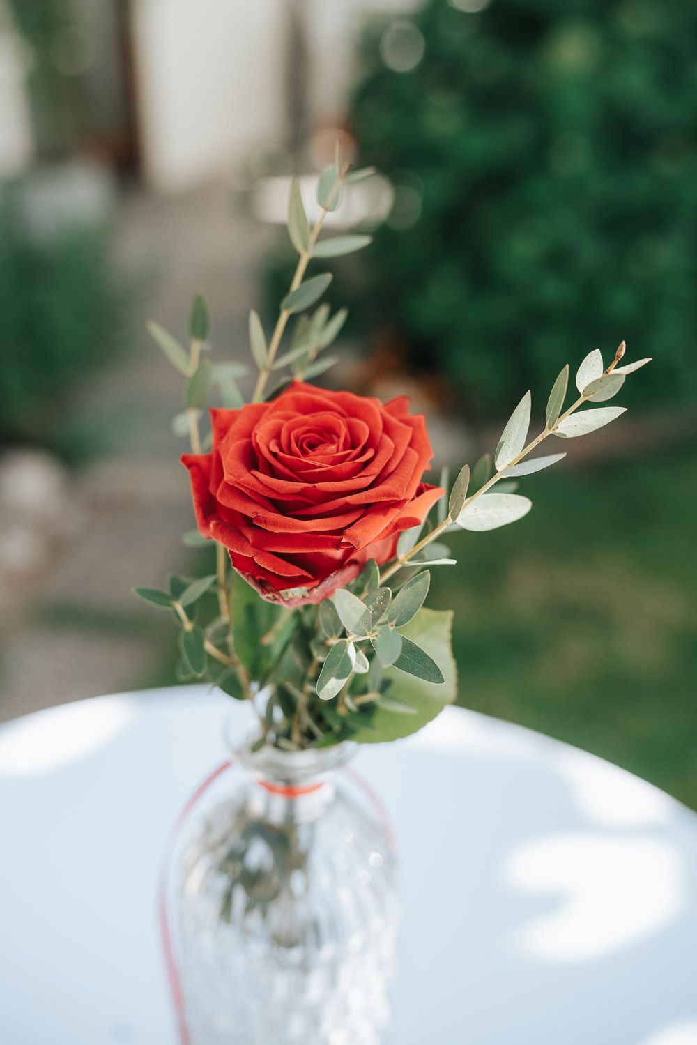 A single red rose with delicate eucalyptus sprigs in a clear glass vase on a white surface.