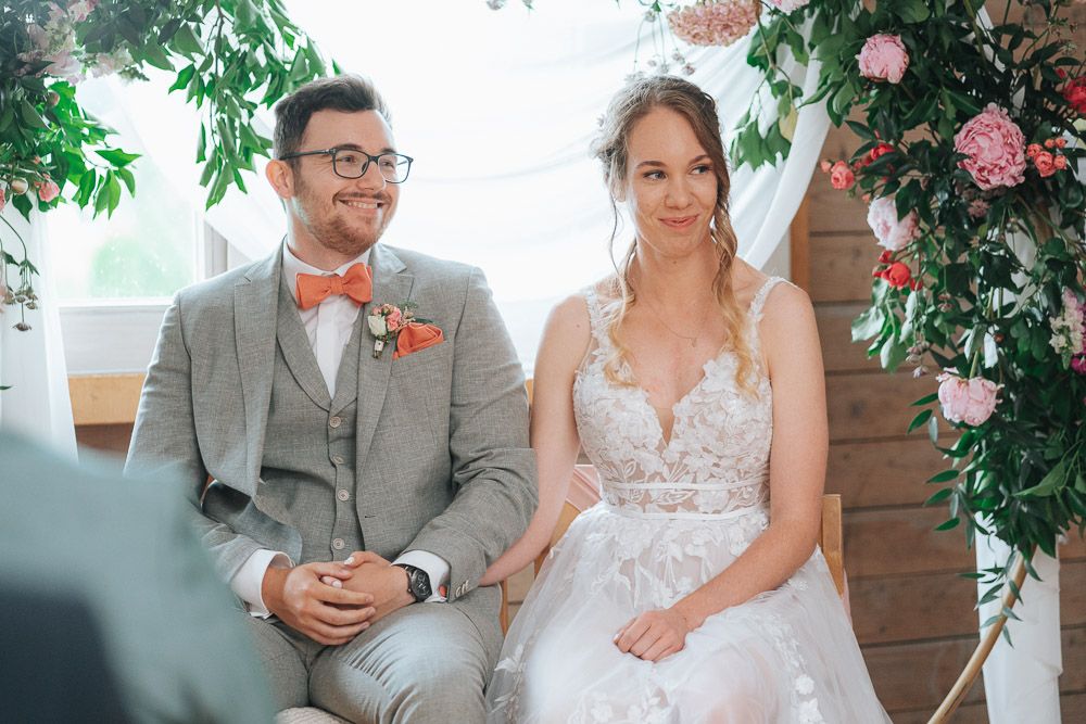 A couple sits together during their wedding ceremony under an arch decorated with pink flowers and greenery.