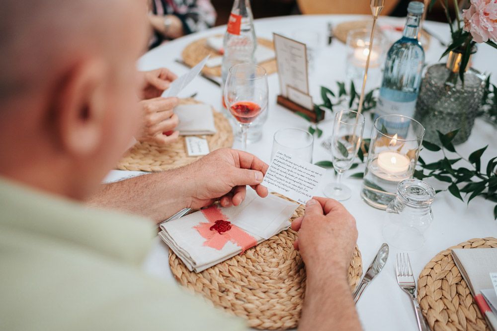 A person's hands arrange a coral-colored place card on a woven placemat at an elegant dinner table with candles and decor.