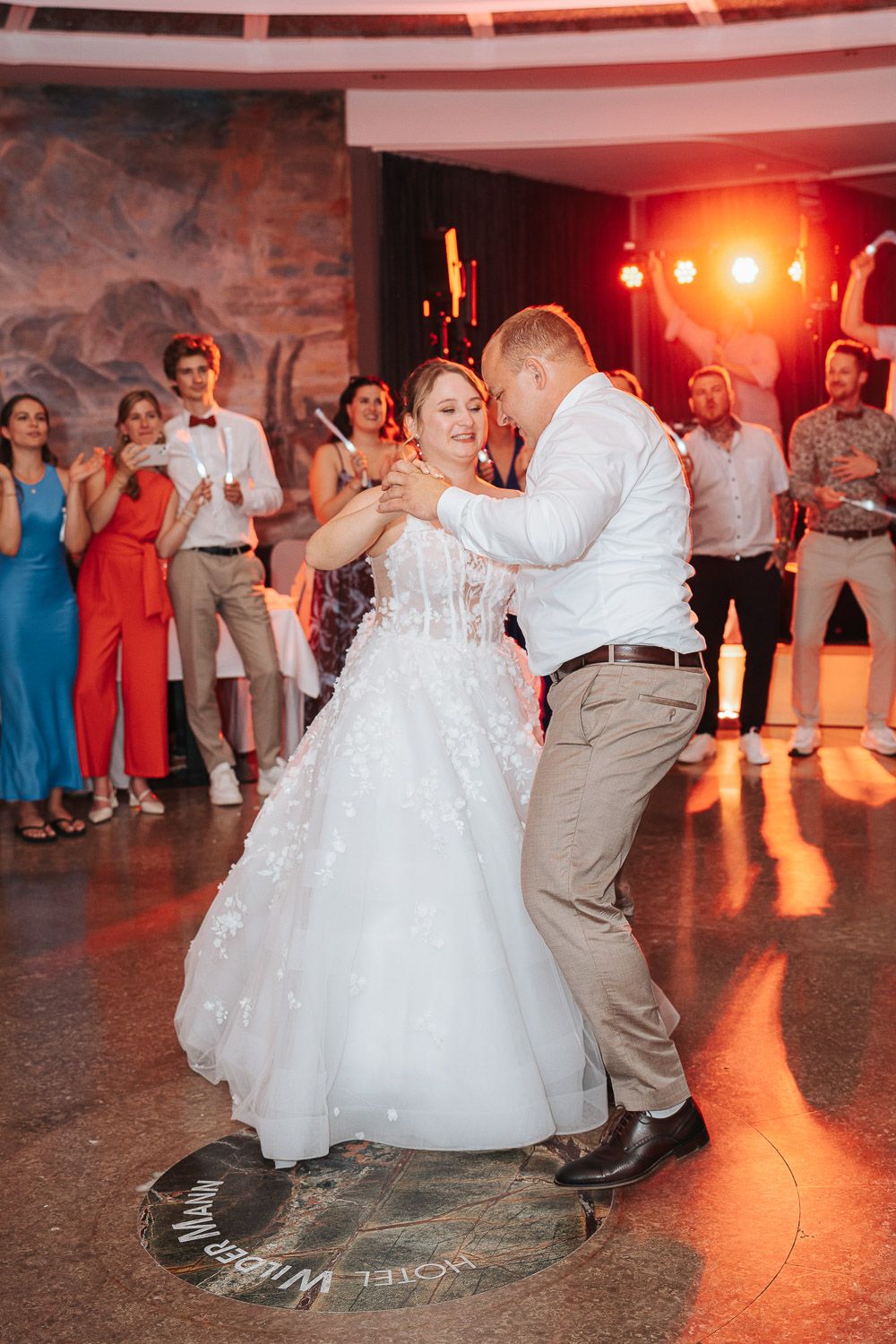 A couple shares their first dance at a wedding reception with guests watching from the dance floor under warm lighting.